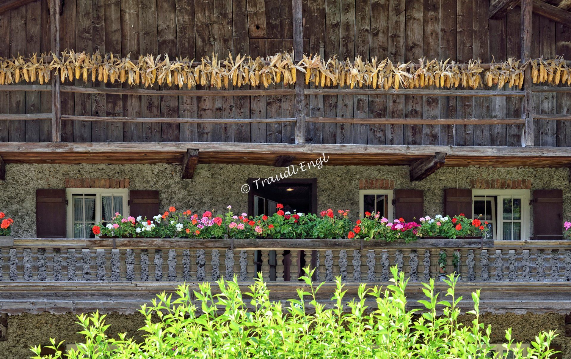 ländliche Hausfassade, Natursteinhaus, Steinhaus, Holzbalkone, Balkonblumen, Blumenschmuck, Maiskolben aufgehängt, Traudl Engl