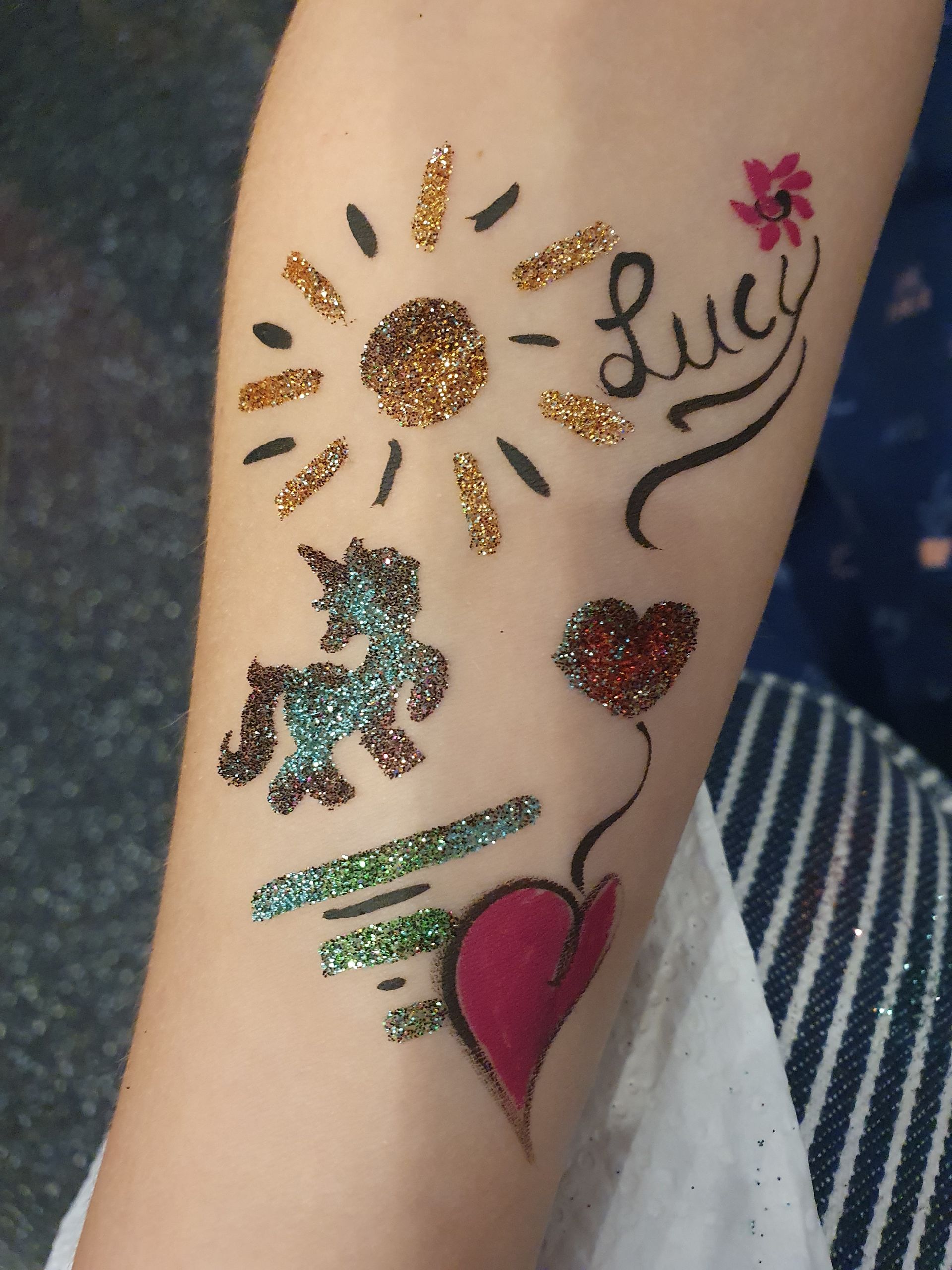 Der Arm ist mit bunten Glitzertattoos bedeckt, darunter eine Sonne, ein Herz, eine Tänzerin und der Name „Lily“.