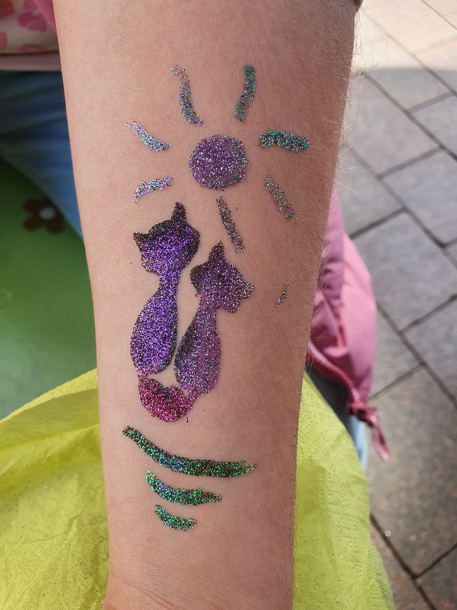Glitzerndes Blumen-Tattoo auf dem Unterarm mit lila Blütenblättern und grünen Blättern.