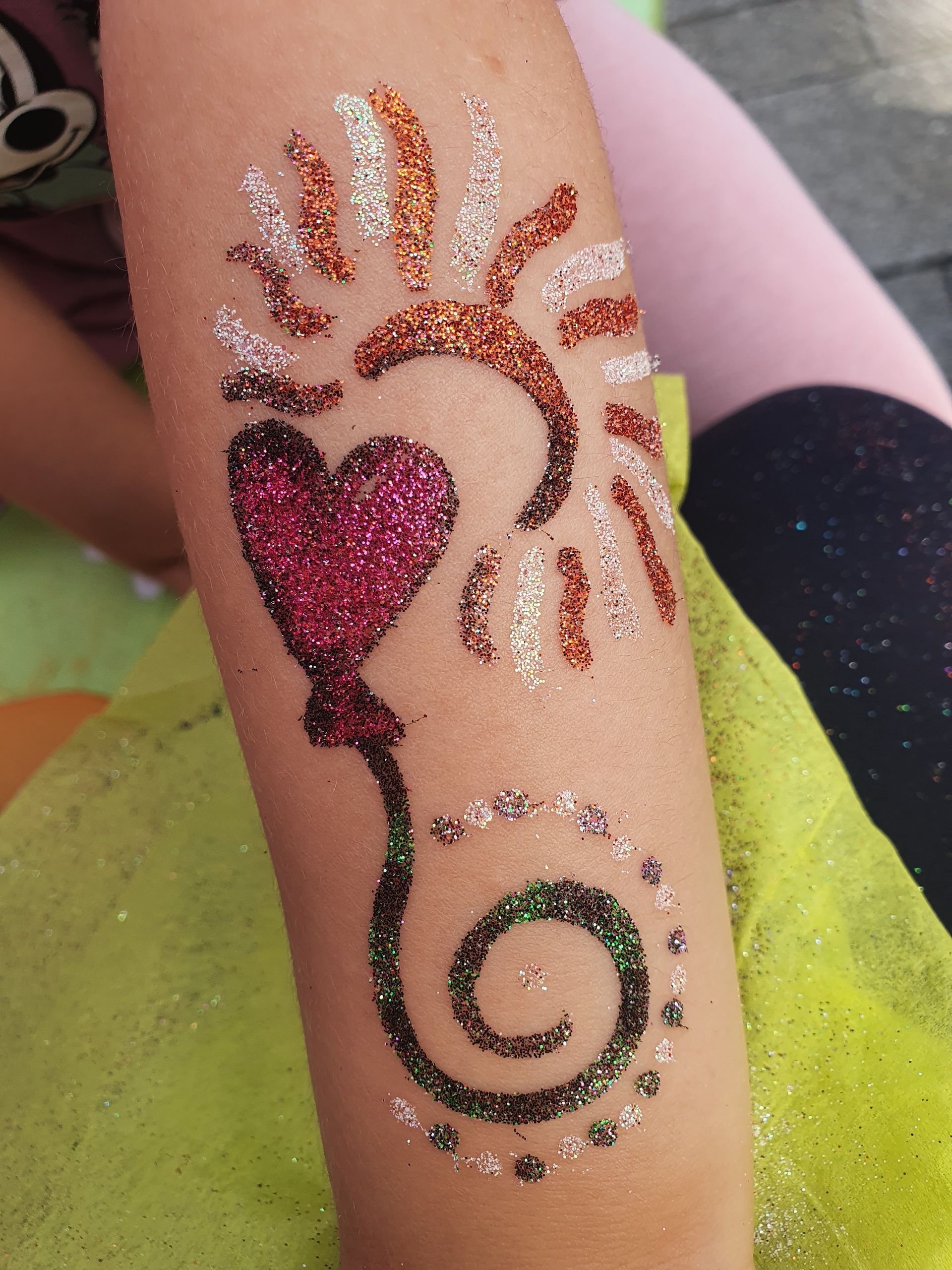 Glitzerndes Henna-Design auf der Haut mit einem rosa Herz, sonnenförmigen Linien und einer grünen Spirale.