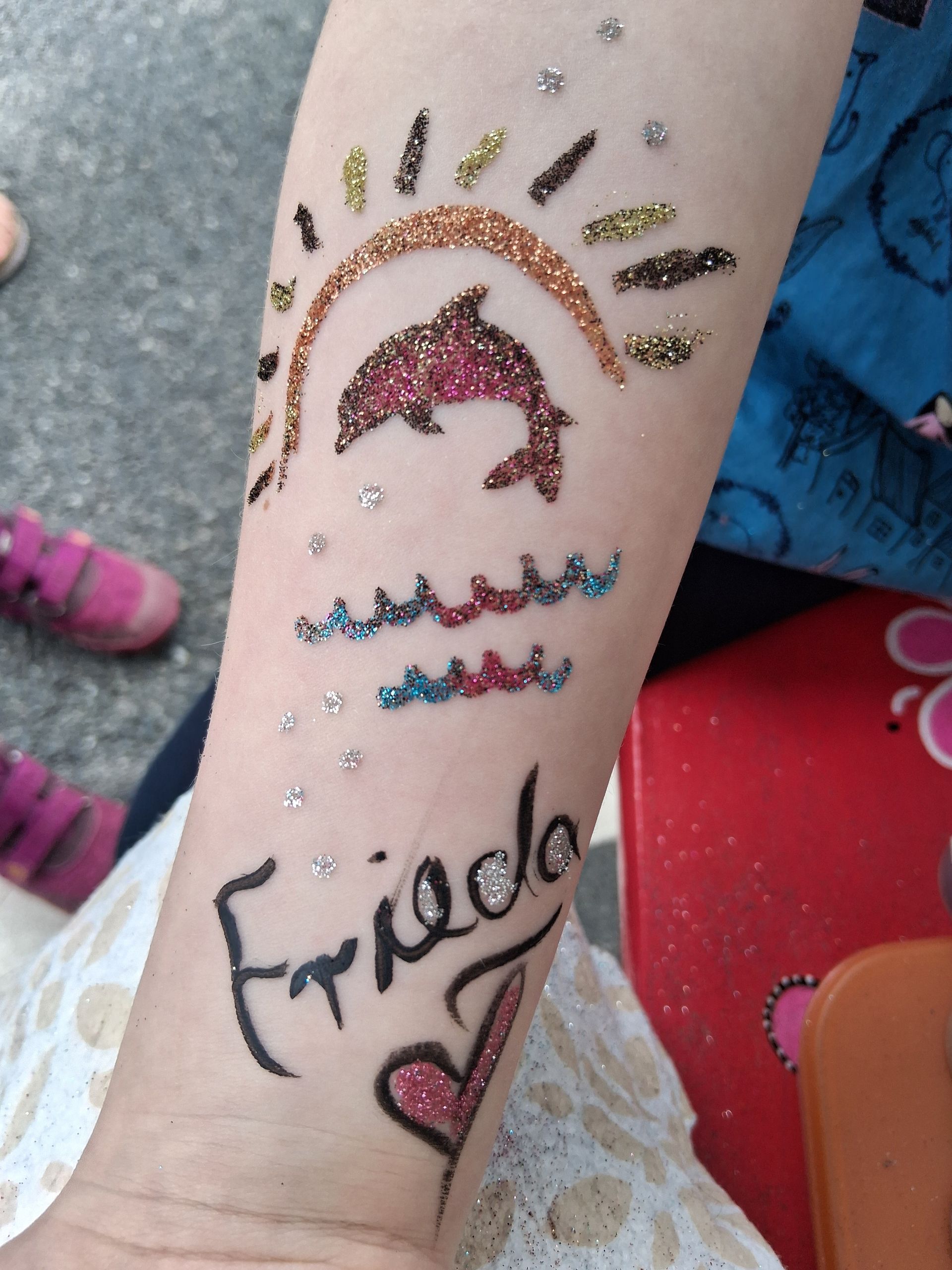 Unterarm mit Glitzertattoos, darunter ein Regenbogen, ein Delfin, Wellen und das Wort „Freund“ mit einem Herz