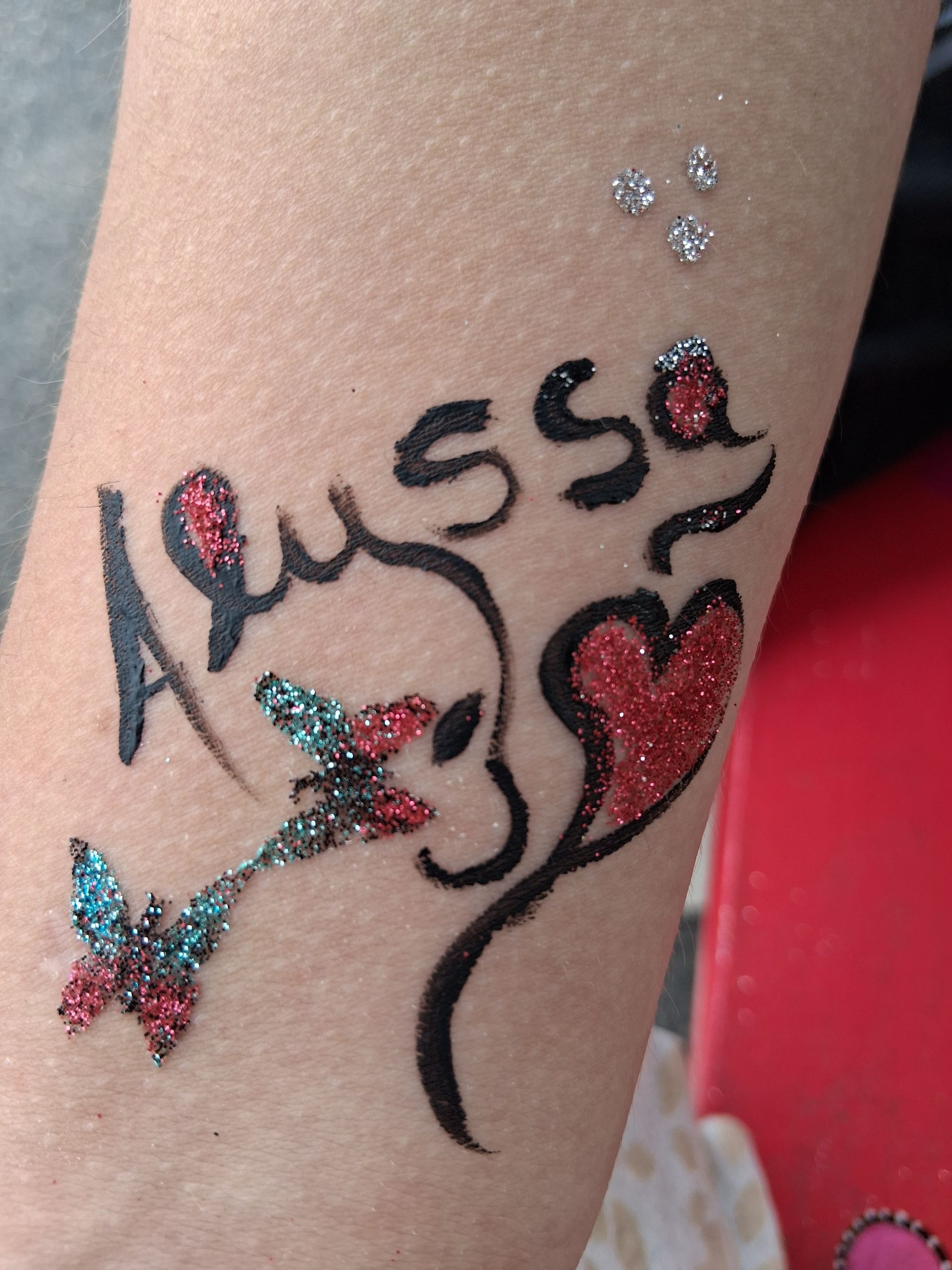 Nahaufnahme von glitzernder Körperkunst mit der Aufschrift „Aussie“ aus Herzen und einem bunten Schmetterling auf der Haut