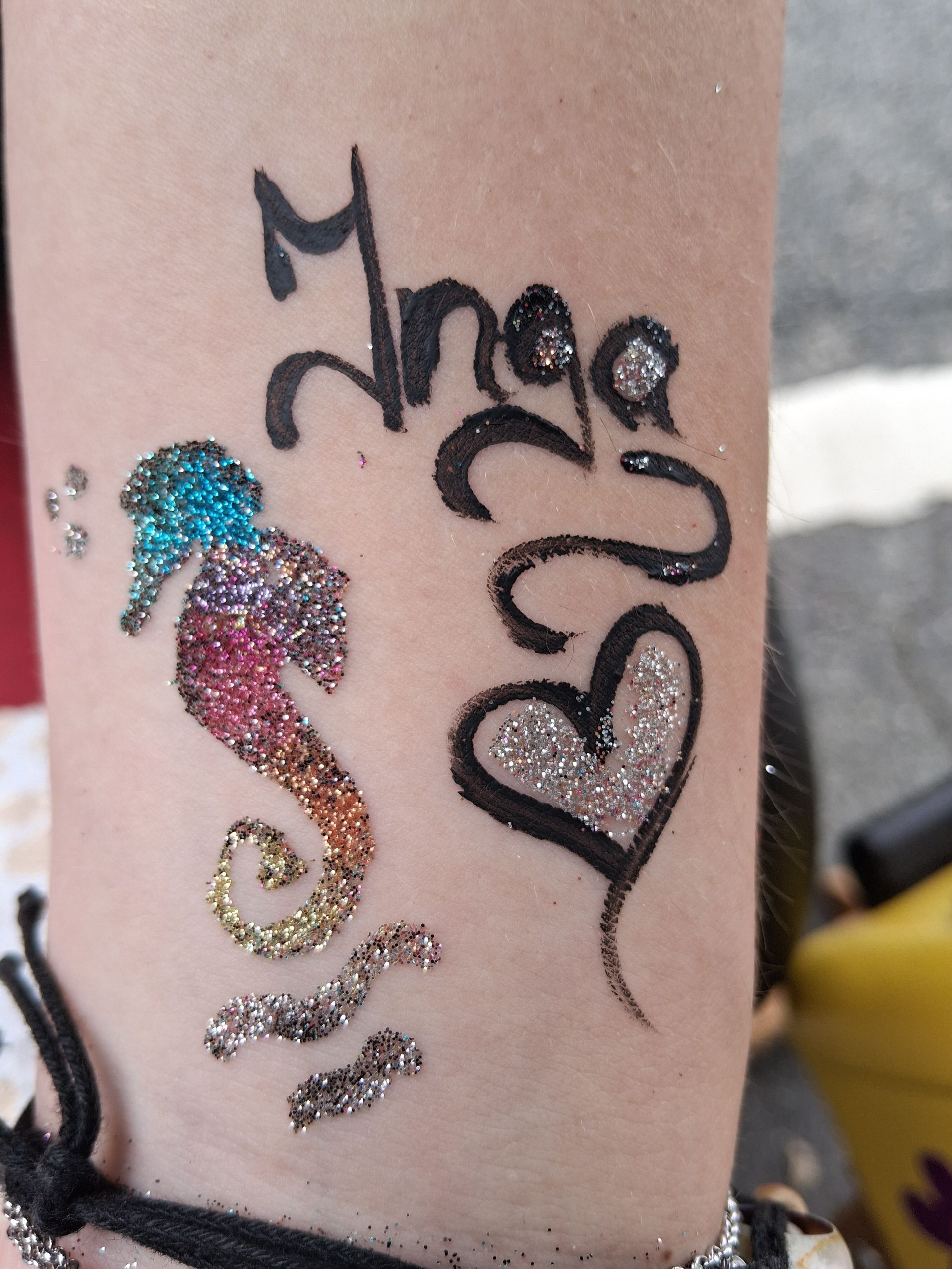 Glitzernde Seepferdchen- und schwarze Herz-Tattoos auf einem Unterarm mit dekorativer Schrift.