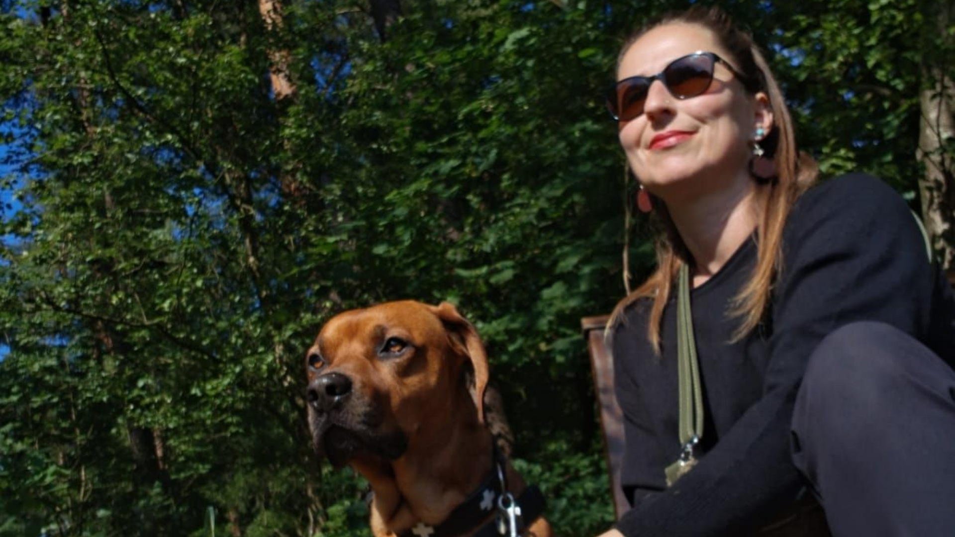 Eine Frau mit Sonnenbrille sitzt im Freien mit einem braunen Hund vor einem grünen Hintergrund.