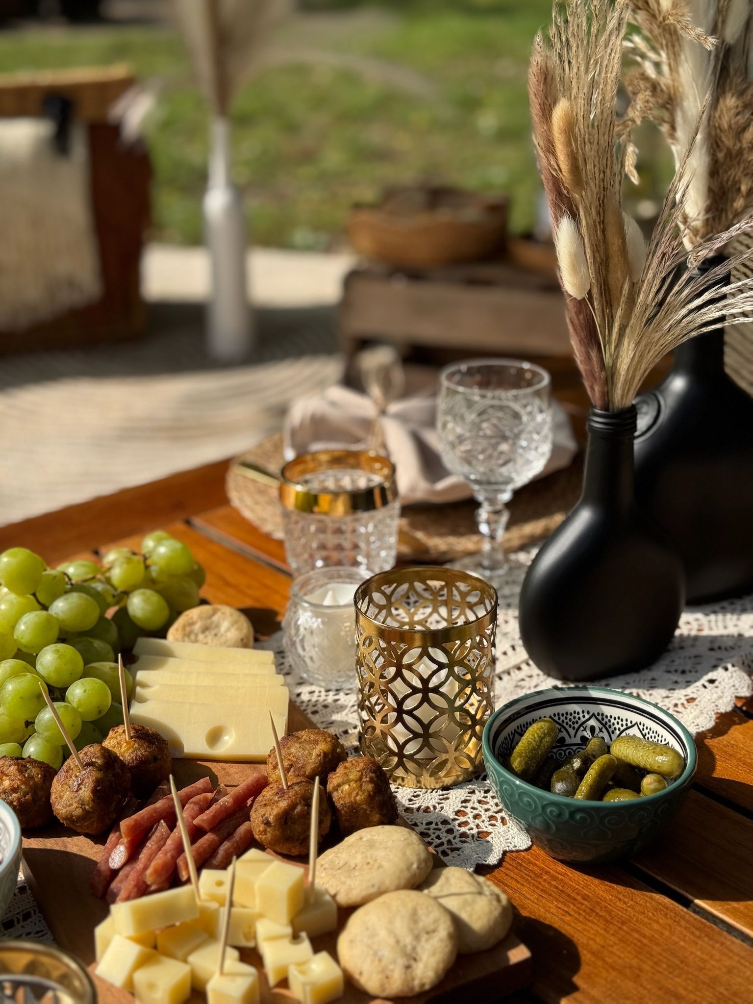 Wurstplatte mit Trauben, Käse und eingelegtem Gemüse, gedeckt für ein Picknick im Freien mit Blumen und Glaswaren.