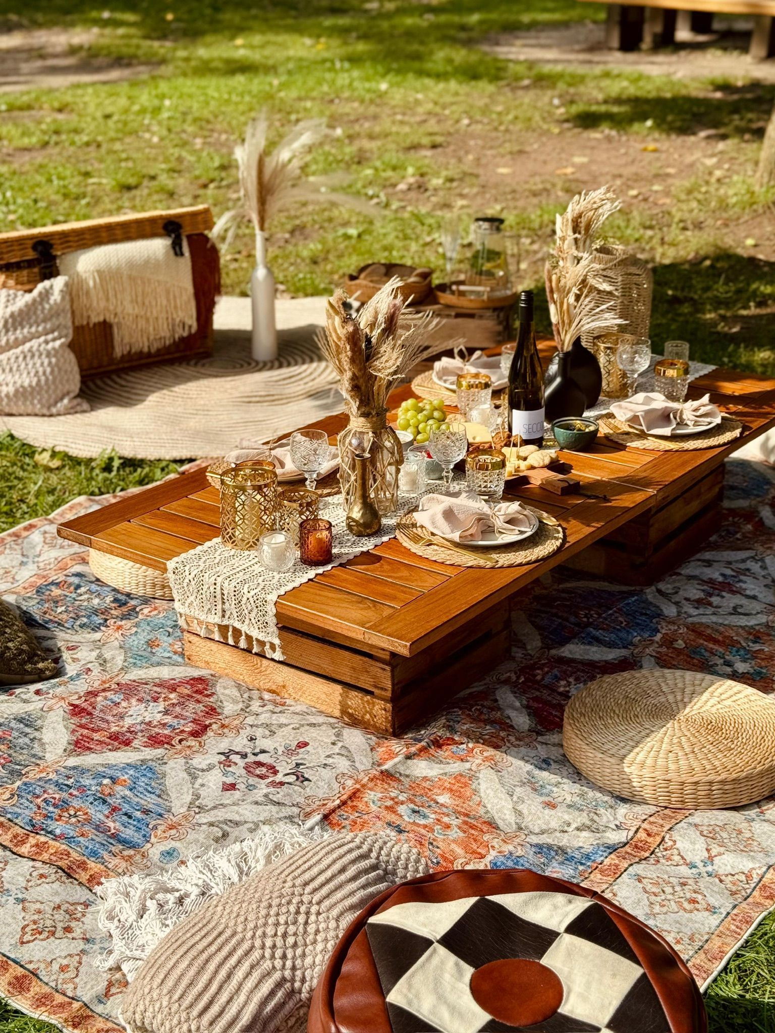 Boho-Picknick auf einem bunt gemusterten Teppich auf einer Rasenfläche. Tisch mit Essen, Getränken und dekorativen Kissen.