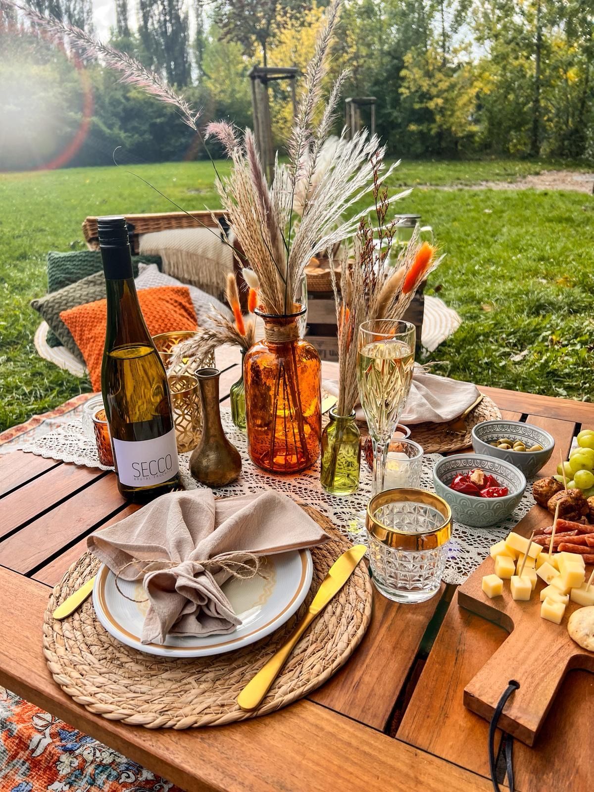 Picknicktisch im Freien mit Essen, Getränken und Blumenschmuck auf Gras.
