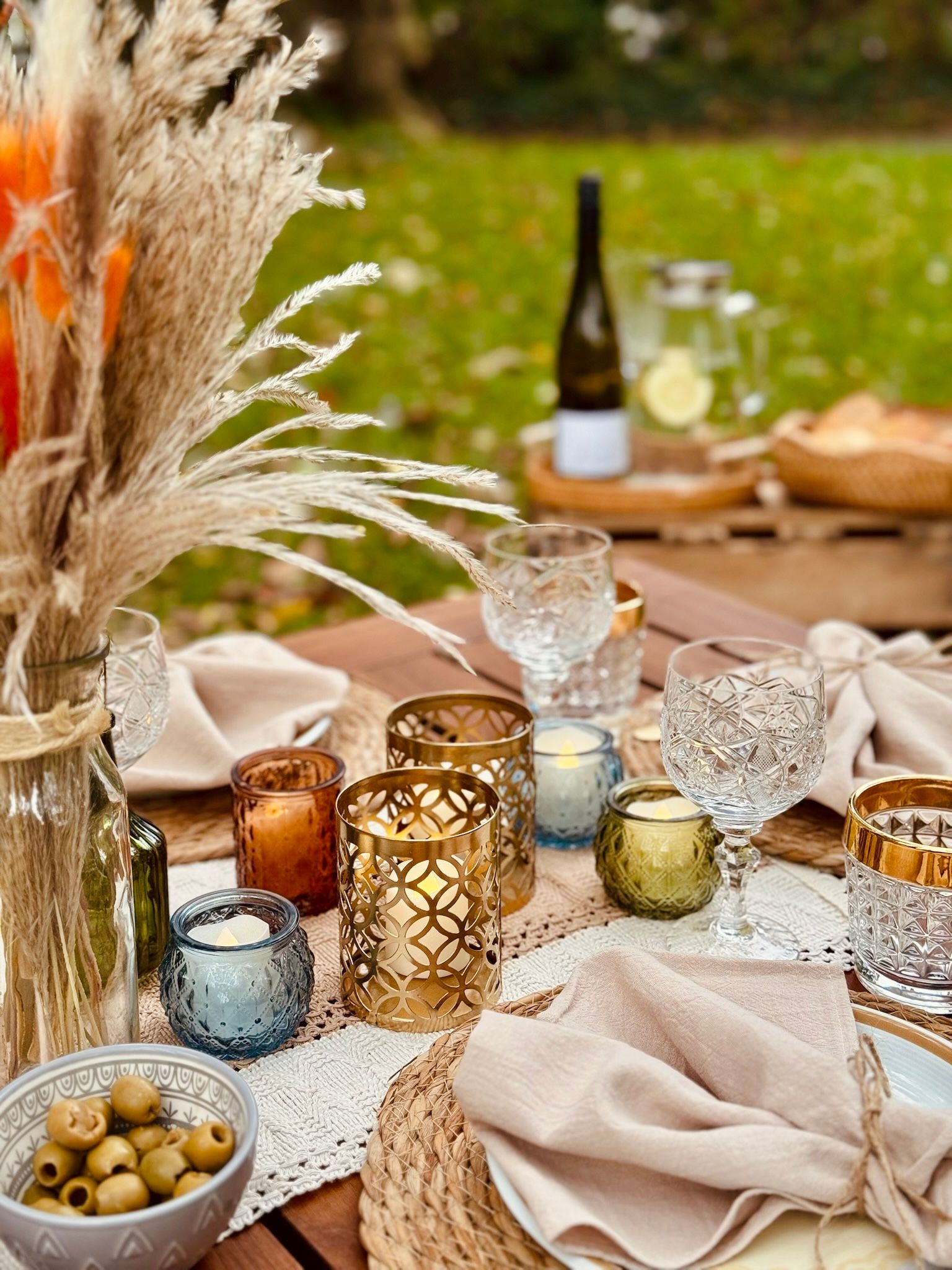 Tischdekoration im Freien mit Kerzen, Gläsern, Servietten, Oliven und einer Flasche Wein.