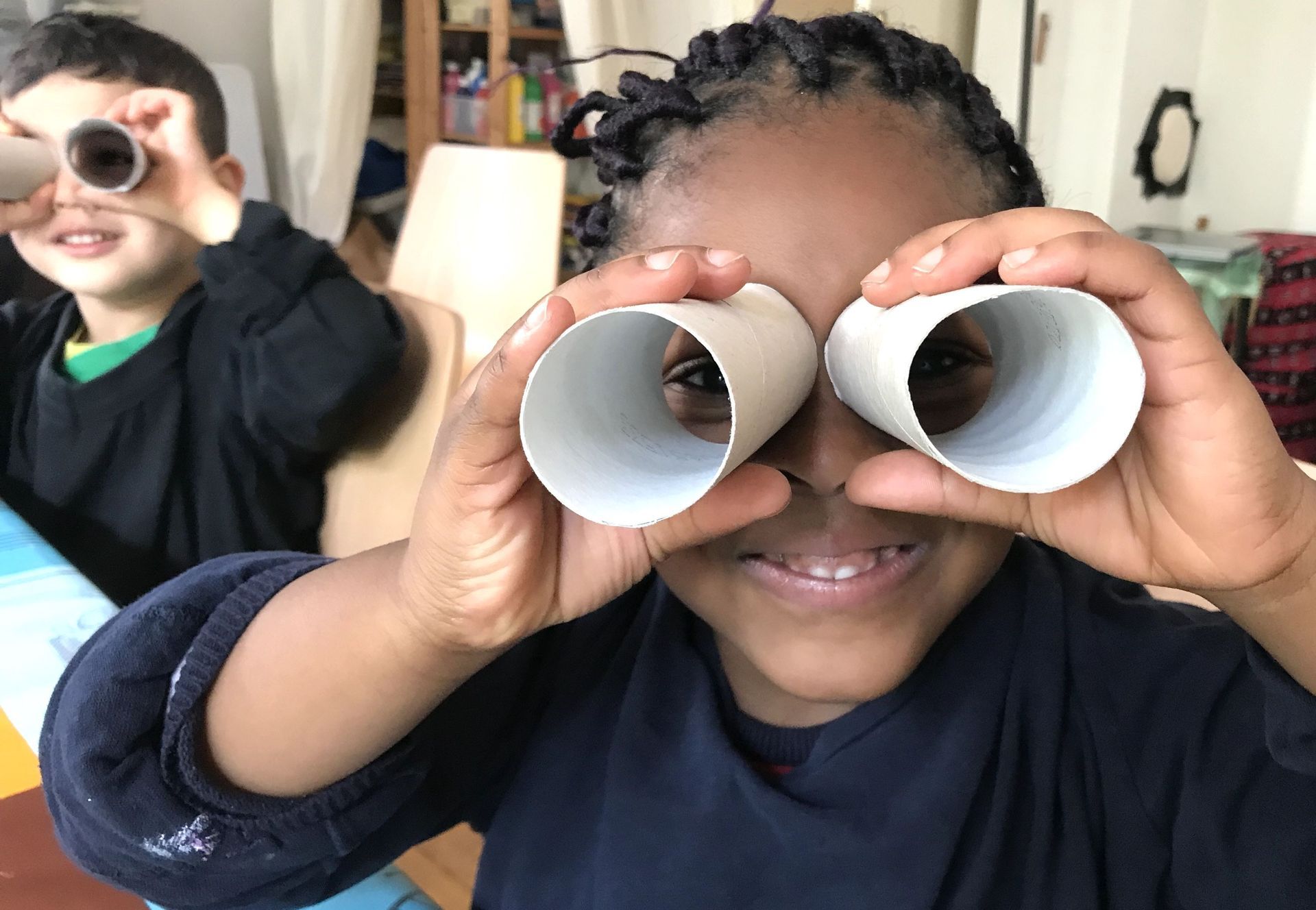Zwei Kinder halten sich drinnen Pappröhren wie Ferngläser vor die Augen.