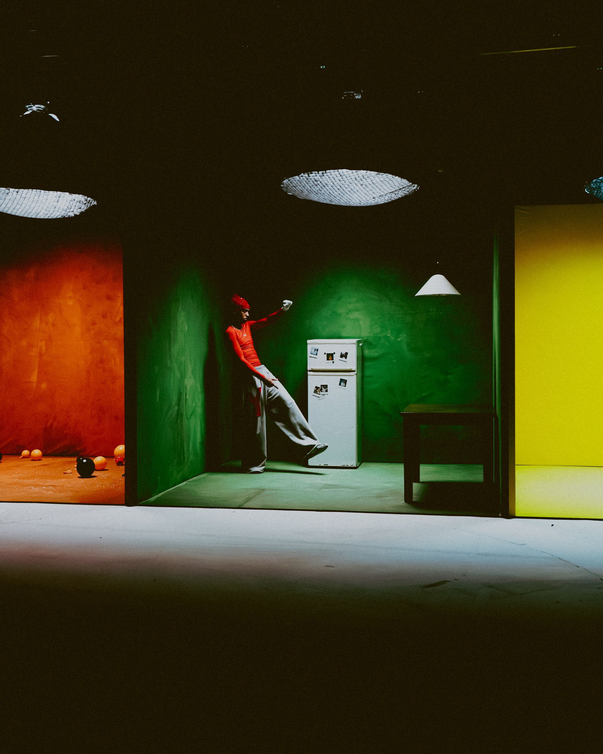 Eine Person in Rot lehnt an einer grünen Wand, zwischen orangefarbenen und gelben Türrahmen. Kühlschrank und Tisch sind vorhanden. Wiz The Mc Videoproduktion Yebo