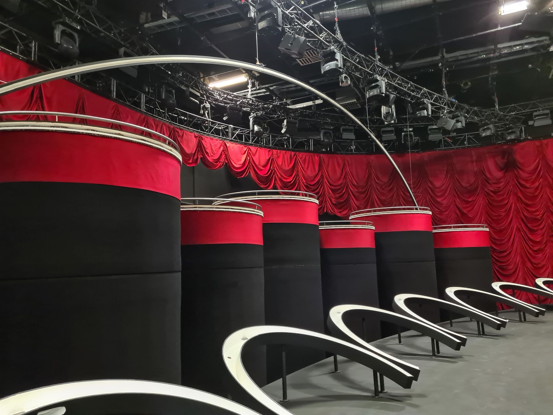 Die Bühne ist mit roten und schwarzen zylindrischen Plattformen, bogenförmigen weißen Konstruktionen und roten Vorhängen ausgestattet.