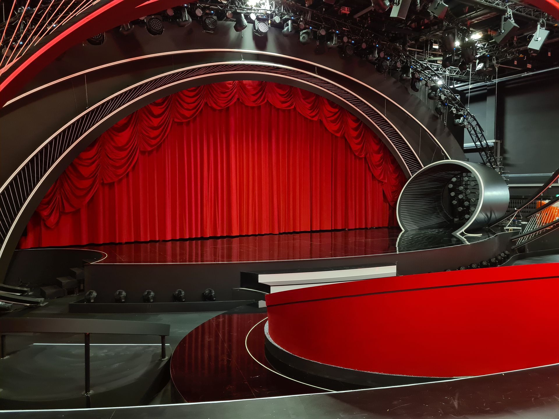 Bühne mit rotem Vorhang, Scheinwerfern und einem gebogenen, roten Podium, bereit für eine Show.