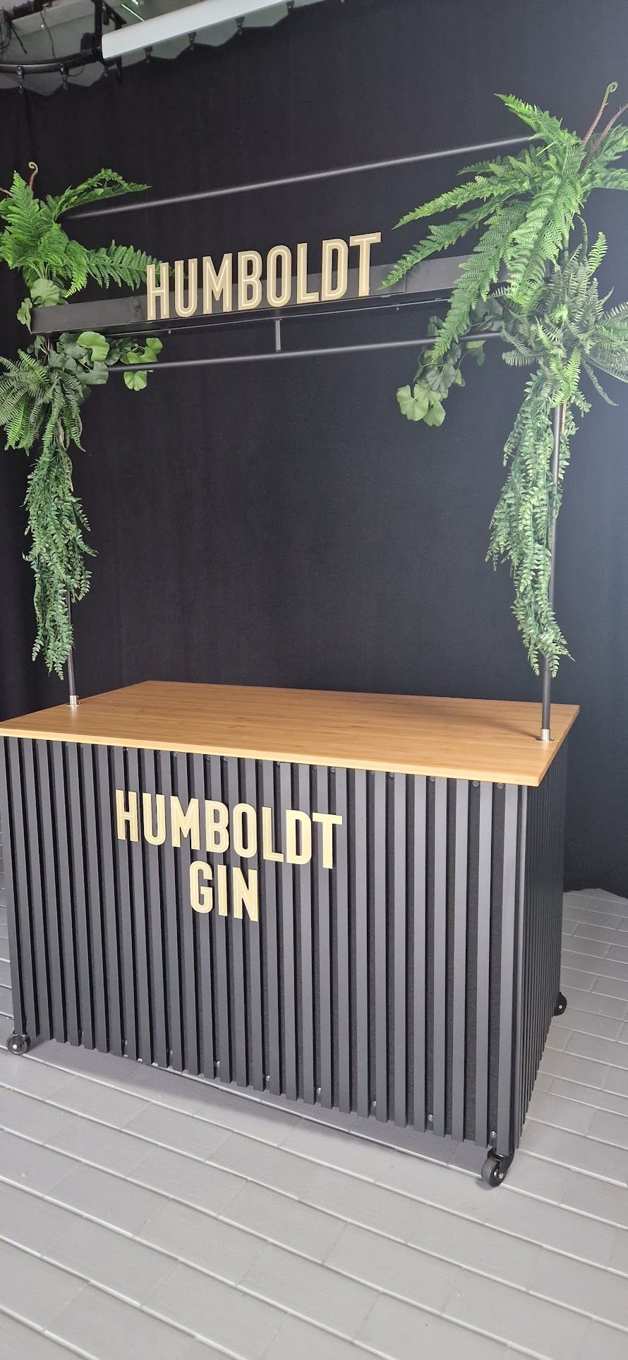 Bar mit „HUMBOLDT GIN“-Schild, Holzplatte, schwarzen Wellblechseiten und Grünpflanzen.