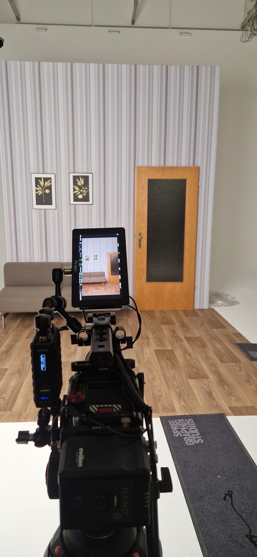 Eine Kamera filmt eine Innenraumszene mit Sofa, Tür und Kunstwerk. Die Aufnahmen entstanden in einem Studio.