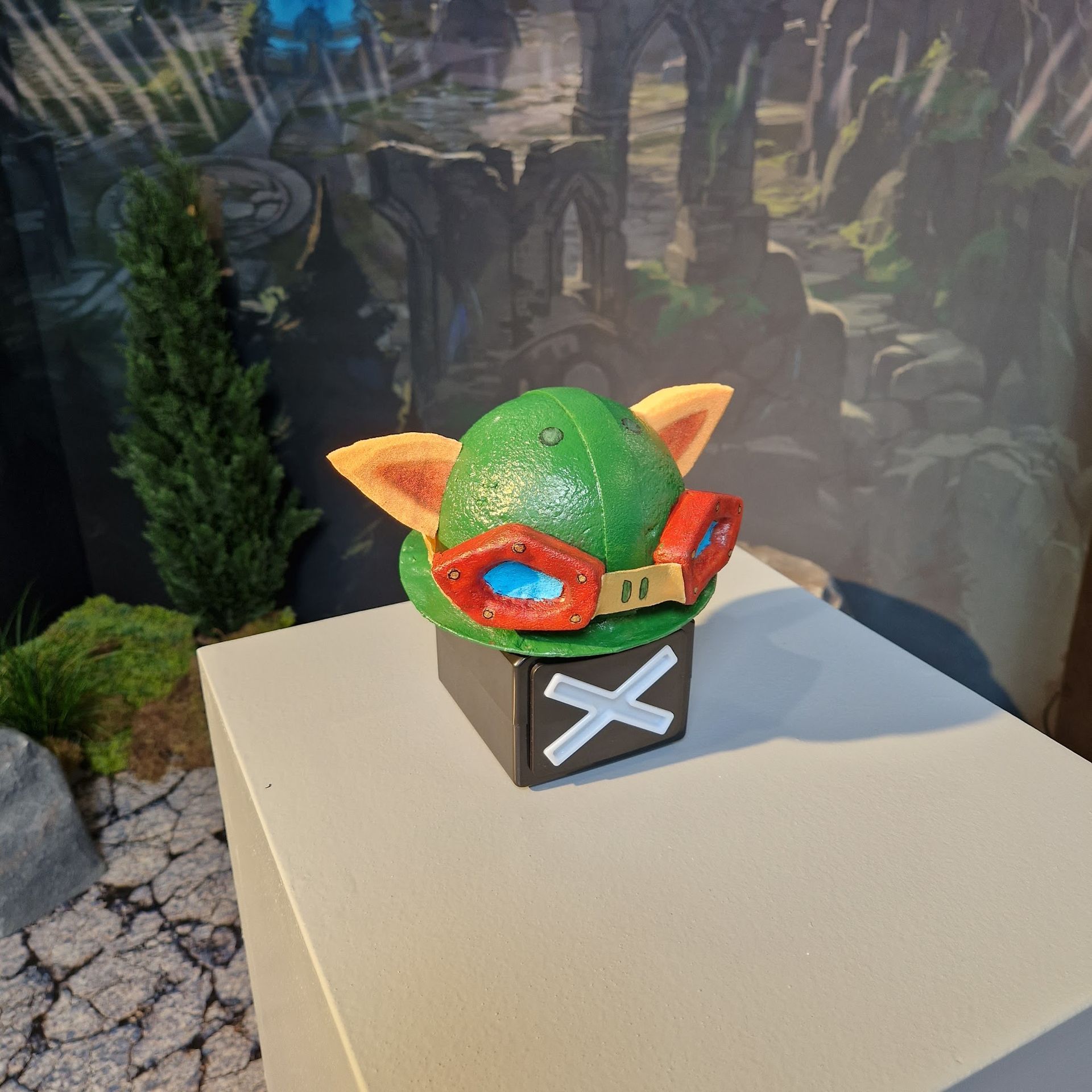 Teemo-Hutskulptur mit Schutzbrille auf einem schwarzen Würfel mit einem weißen „X“ vor einer Waldkulisse.