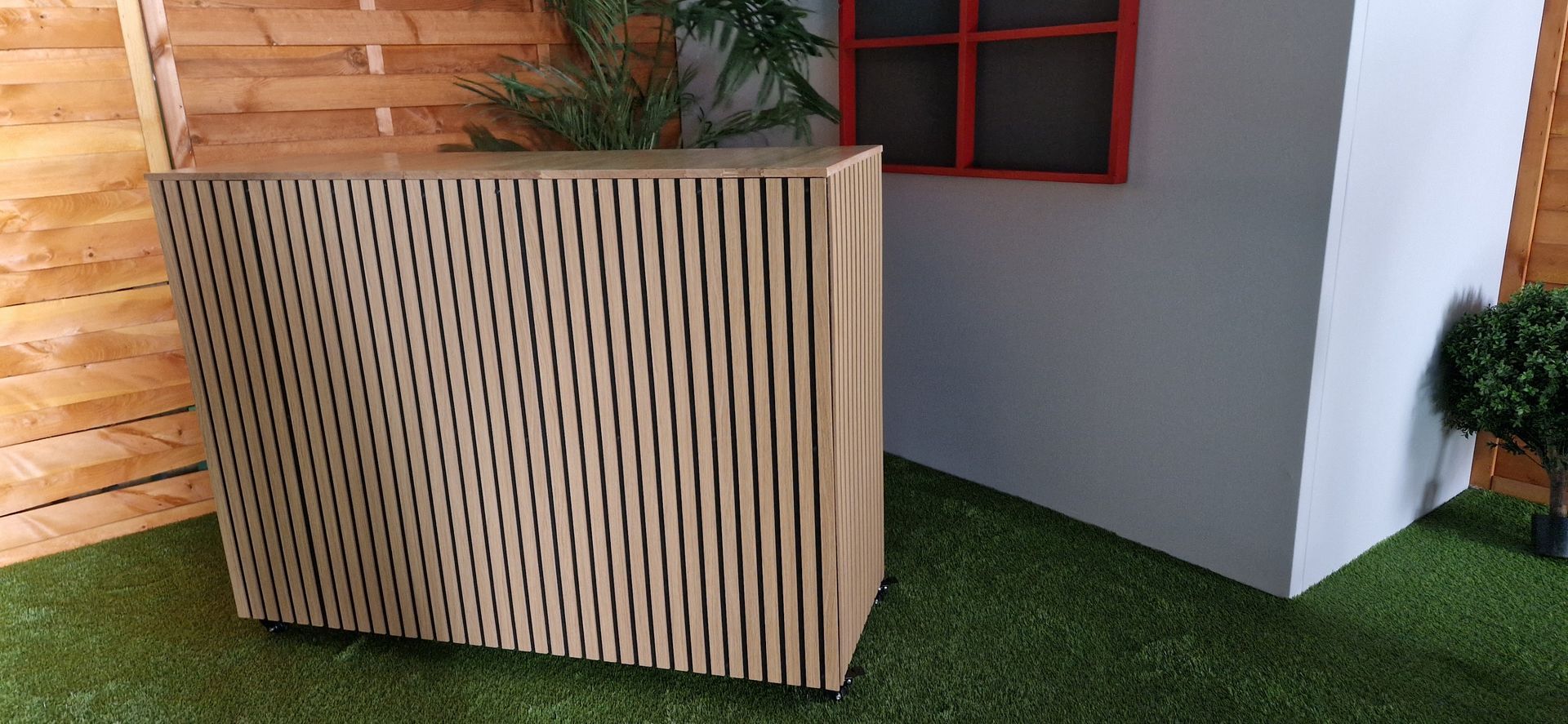 Eine Lamellentheke mit vertikalen Latten steht auf grünem Gras neben einer weißen Wand mit einem kleinen roten Fenster.