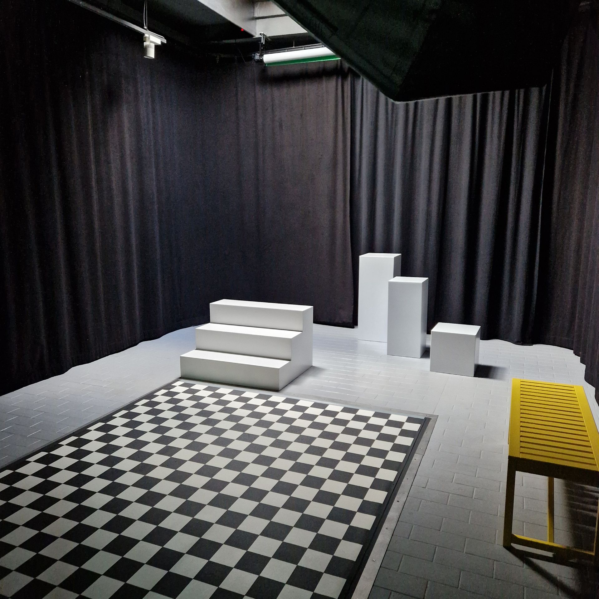 Studio-Set mit schwarzen Vorhängen, Schachbrettboden, weißen Podesten und einem gelben Stuhl.