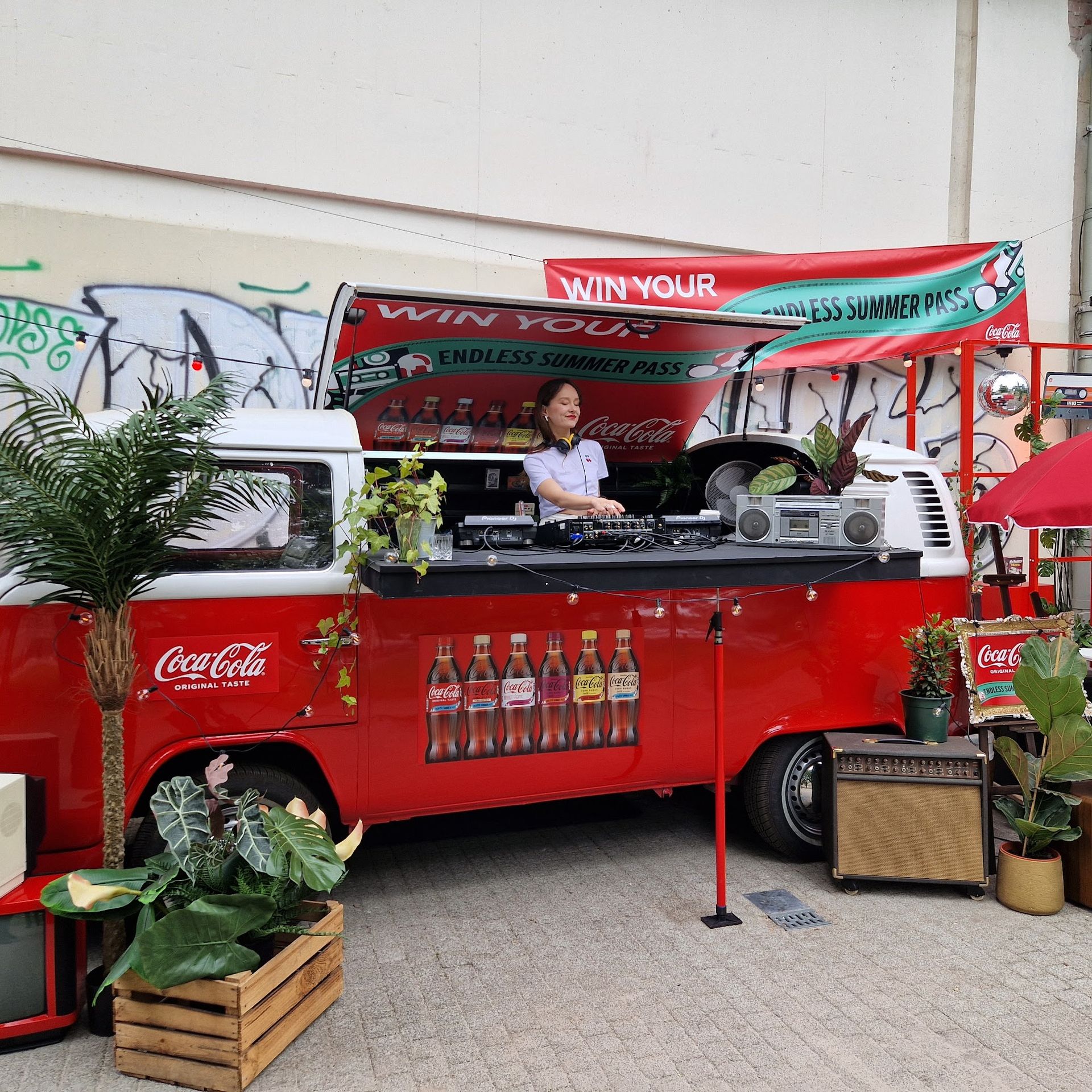 DJ-Pult in einem rot-weißen VW-Bus, Frau an den Bedienelementen. Pflanzen und Coca-Cola-Logos.