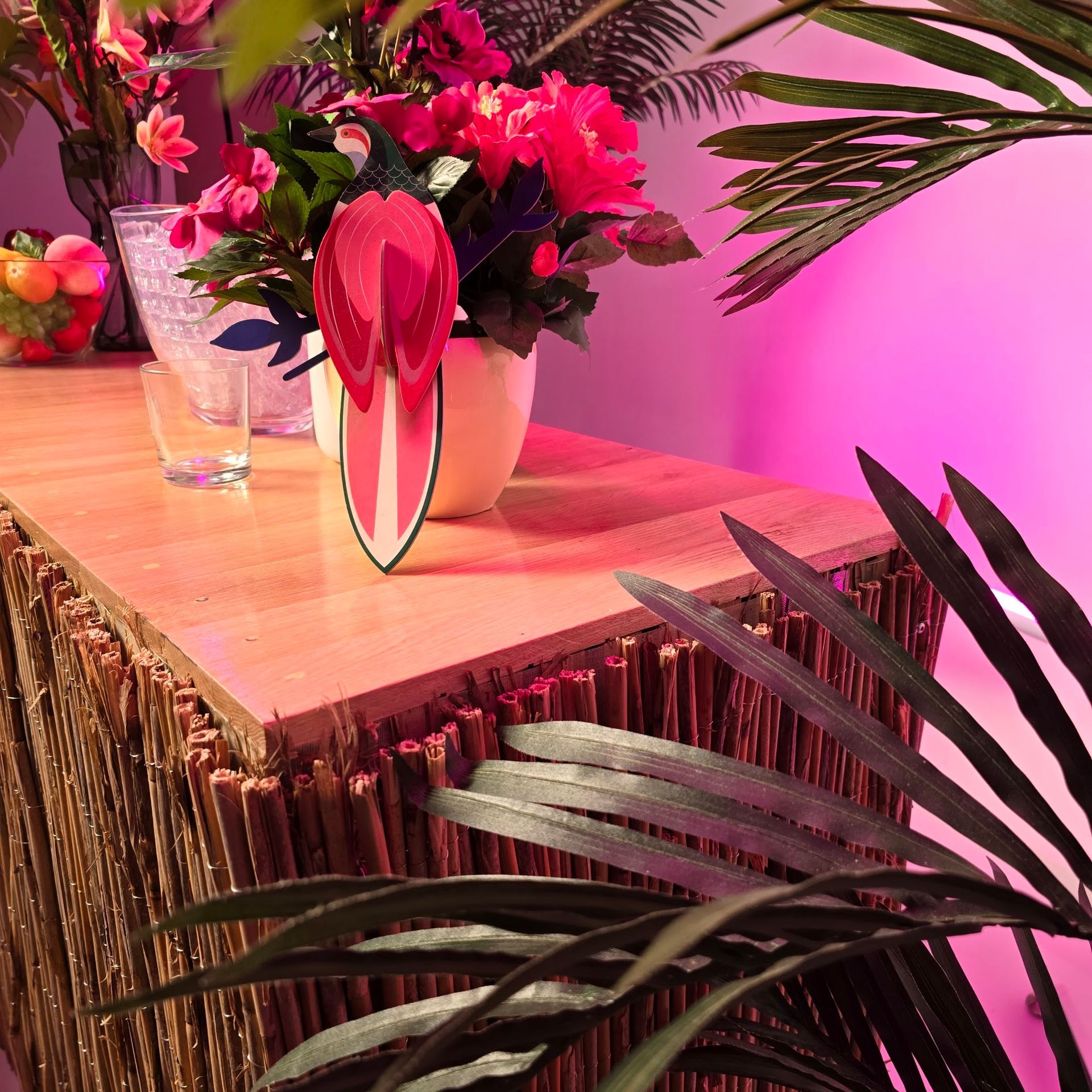 Holztisch mit Blumen, Papierfiguren und Palmenwedeln. Rosa Beleuchtung.