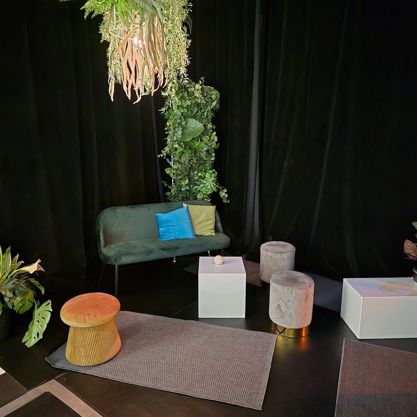 Gemütlicher Loungebereich mit grünem Samtsofa, Zierkissen und Pflanzendekoration vor schwarzen Vorhängen.