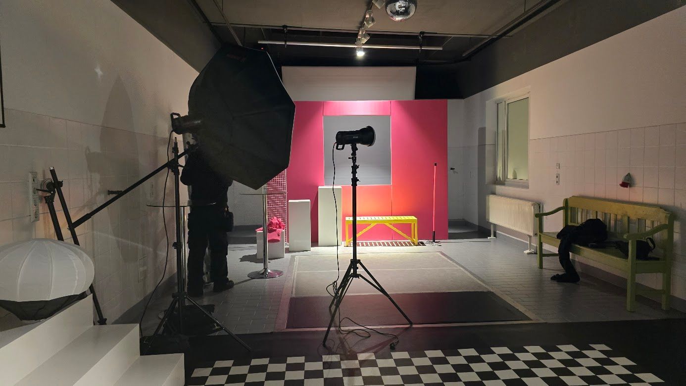 Fotostudio-Aufbau: Softbox und Lampenstative beleuchten einen rosa Hintergrund mit einer gelben Bank. Schachbrettmuster auf dem Boden.