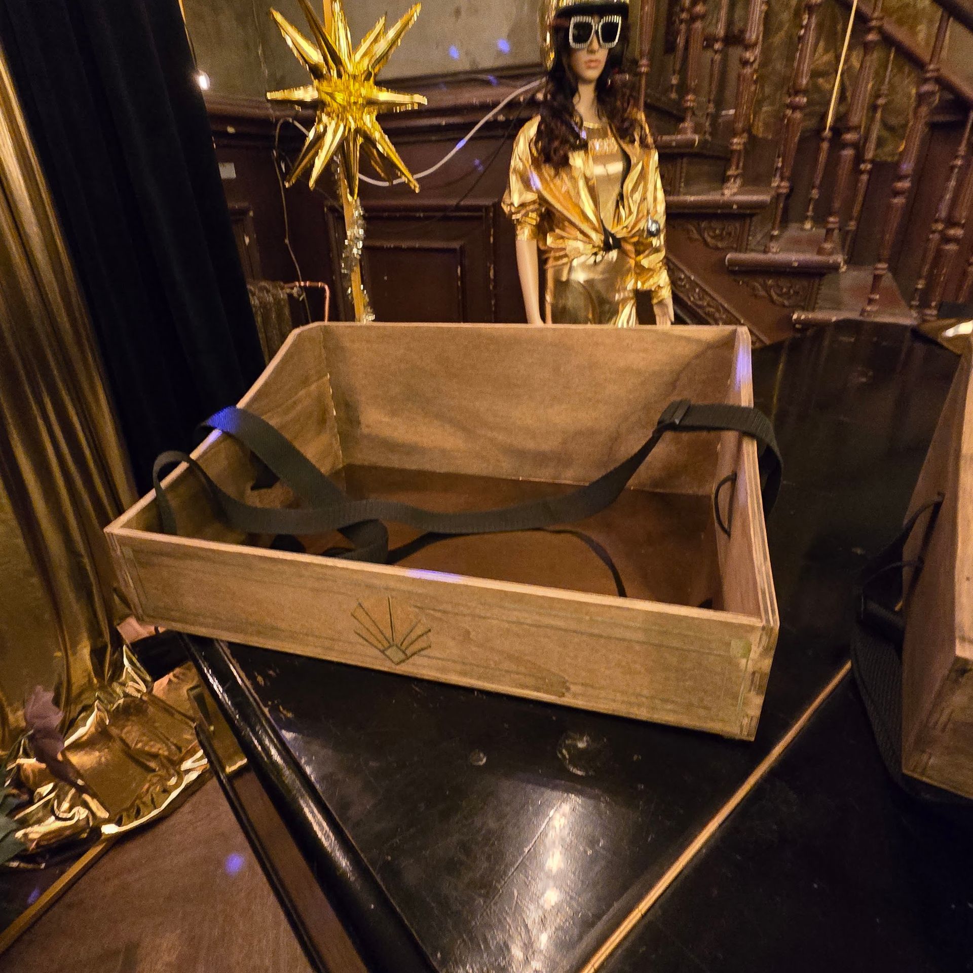 Eine Holzkiste mit Riemen steht auf einem Tisch, im Hintergrund ist ein goldenes Outfit auf einer Schaufensterpuppe zu sehen.