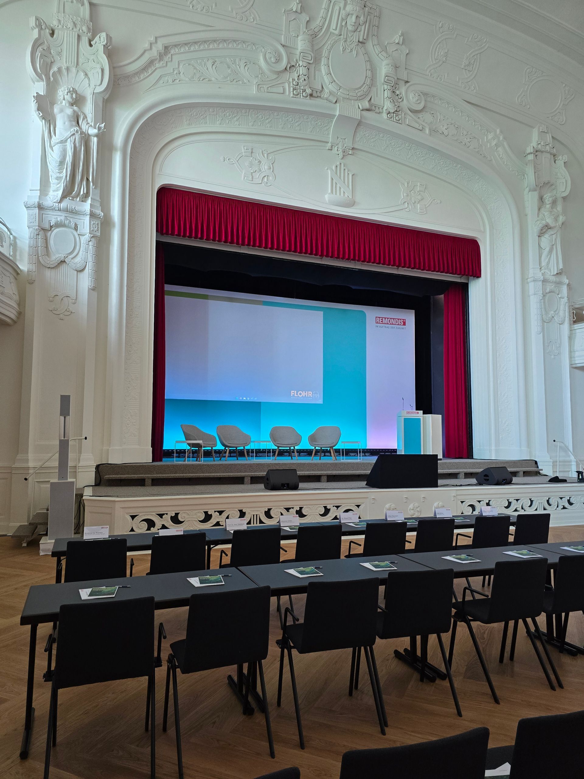 Die Zuschauerplätze sind auf eine Bühne mit Leinwand, Stühlen und Rednerpult in einem weiß getünchten Raum mit kunstvollen Schnitzereien ausgerichtet.