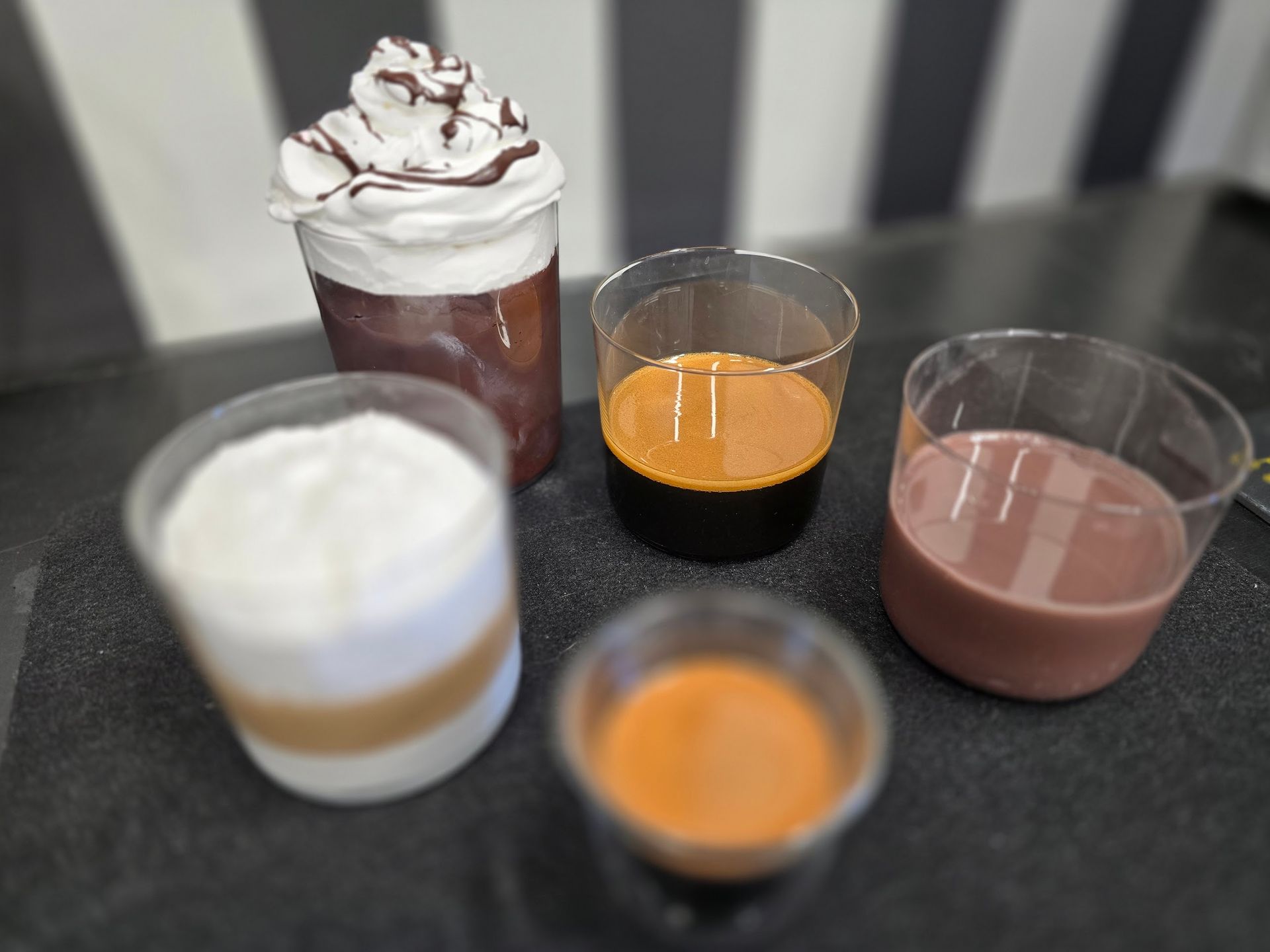 Fünf verschiedene Kaffeegetränke in durchsichtigen Bechern, darunter auch mit Schlagsahne und in verschiedenen Farben.