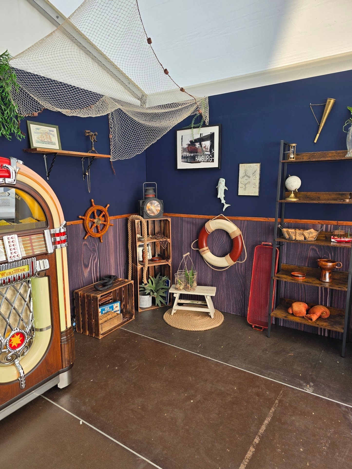 Zimmer im maritimen Stil mit Jukebox, Regalen mit Dekoration, marineblauen und holzgetäfelten Wänden und einer Decke aus Fischernetzen.