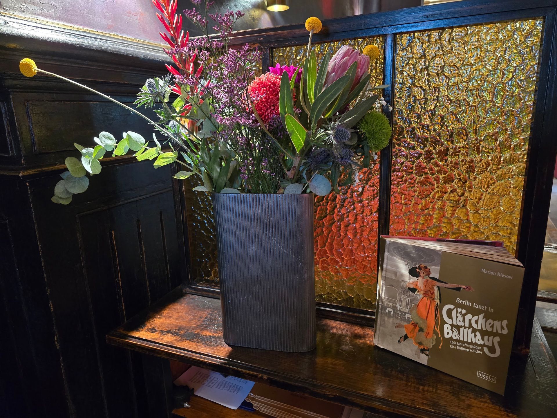 Eine Vase mit bunten Blumen auf einem Regal mit Buntglasfenstern und einem Buch.