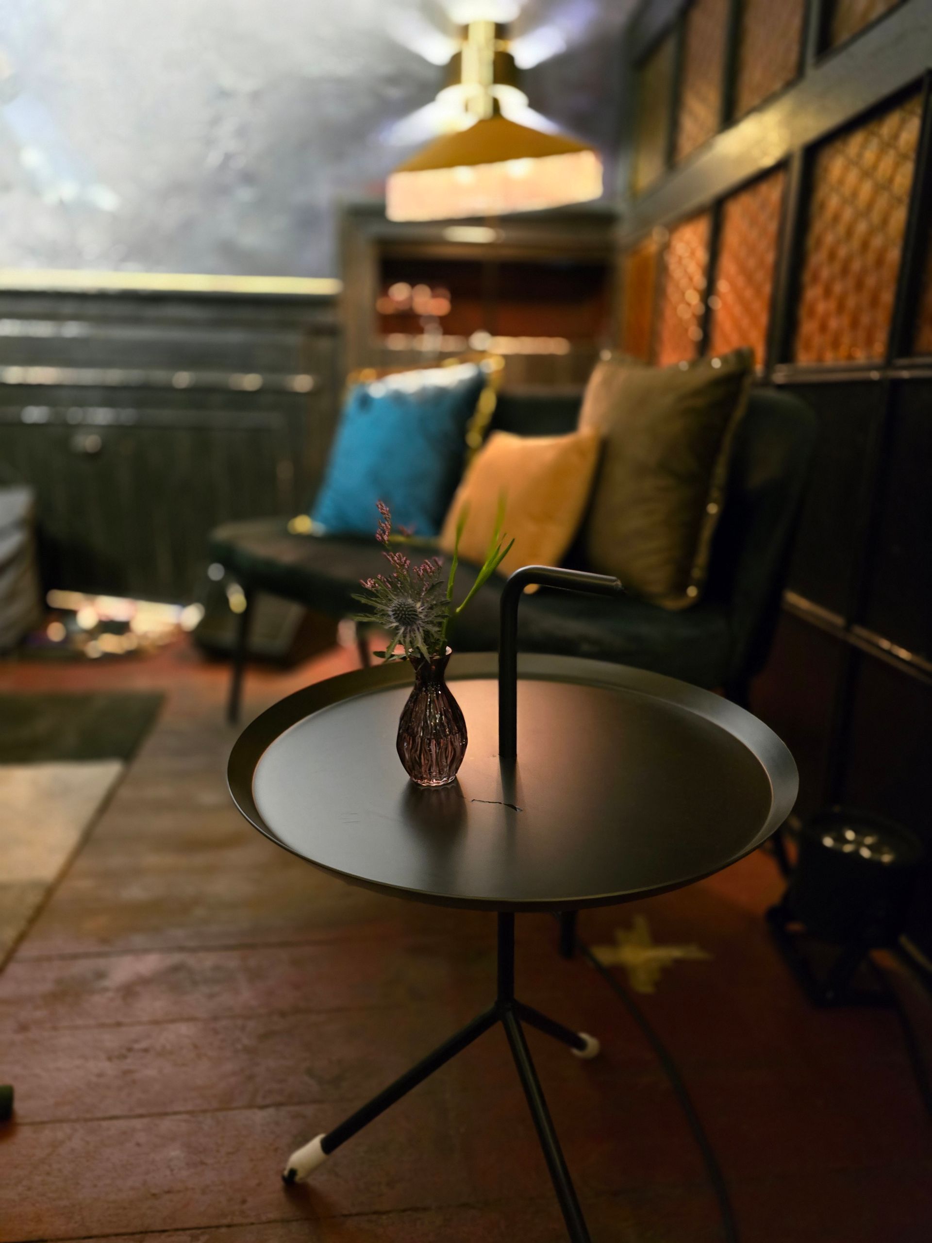 Schwarzer Beistelltisch mit einer Blumenvase in einem schwach beleuchteten Raum mit Sofa und Wandpaneelen.