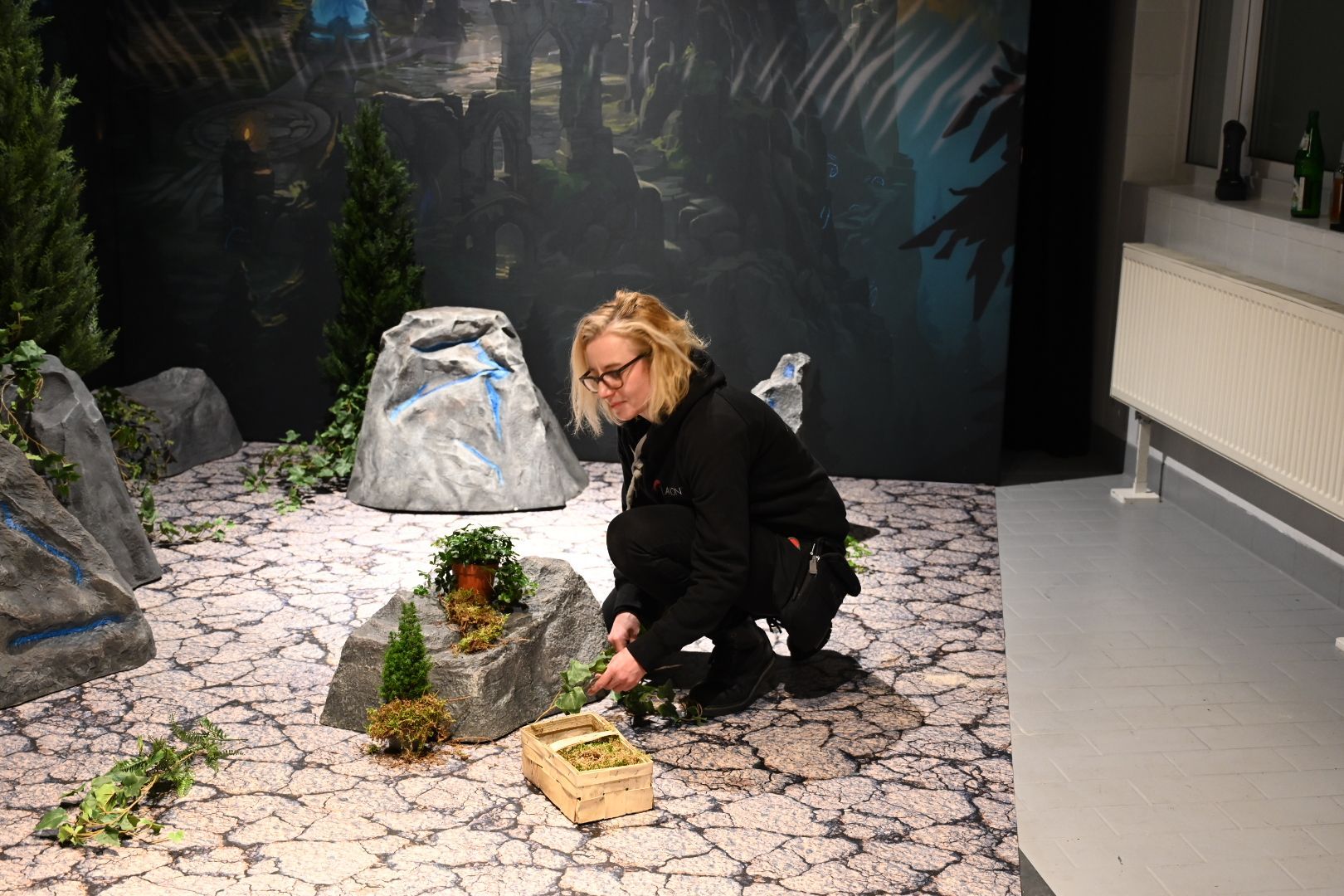 Eine Frau gestaltet eine Miniaturlandschaft mit Felsen, Bäumen und einem Wandgemälde im Hintergrund.