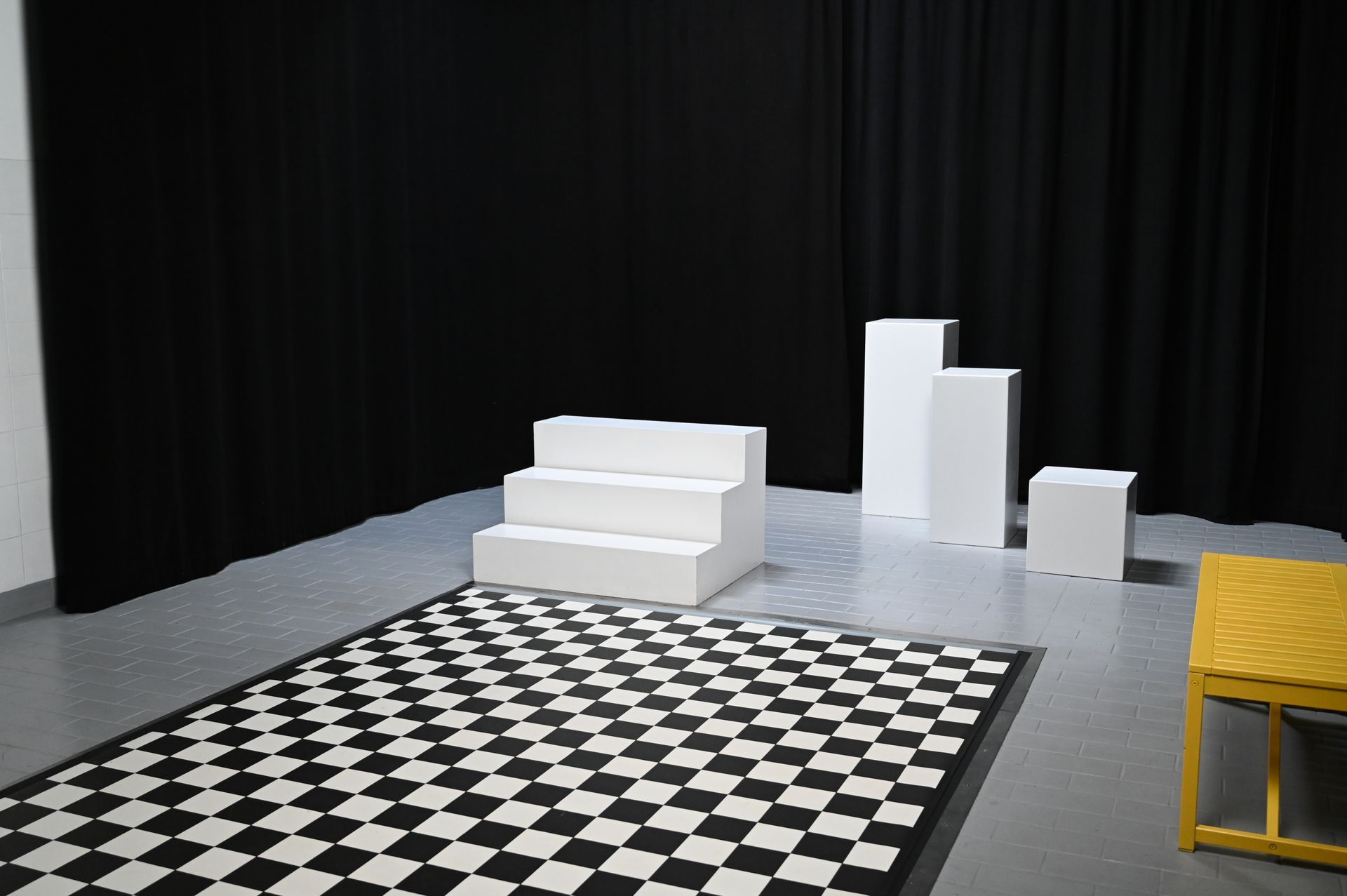 Ein Studio mit einem karierten Teppich, weißen Podesten und schwarzen Vorhängen. An der Seite steht ein gelber Stuhl.