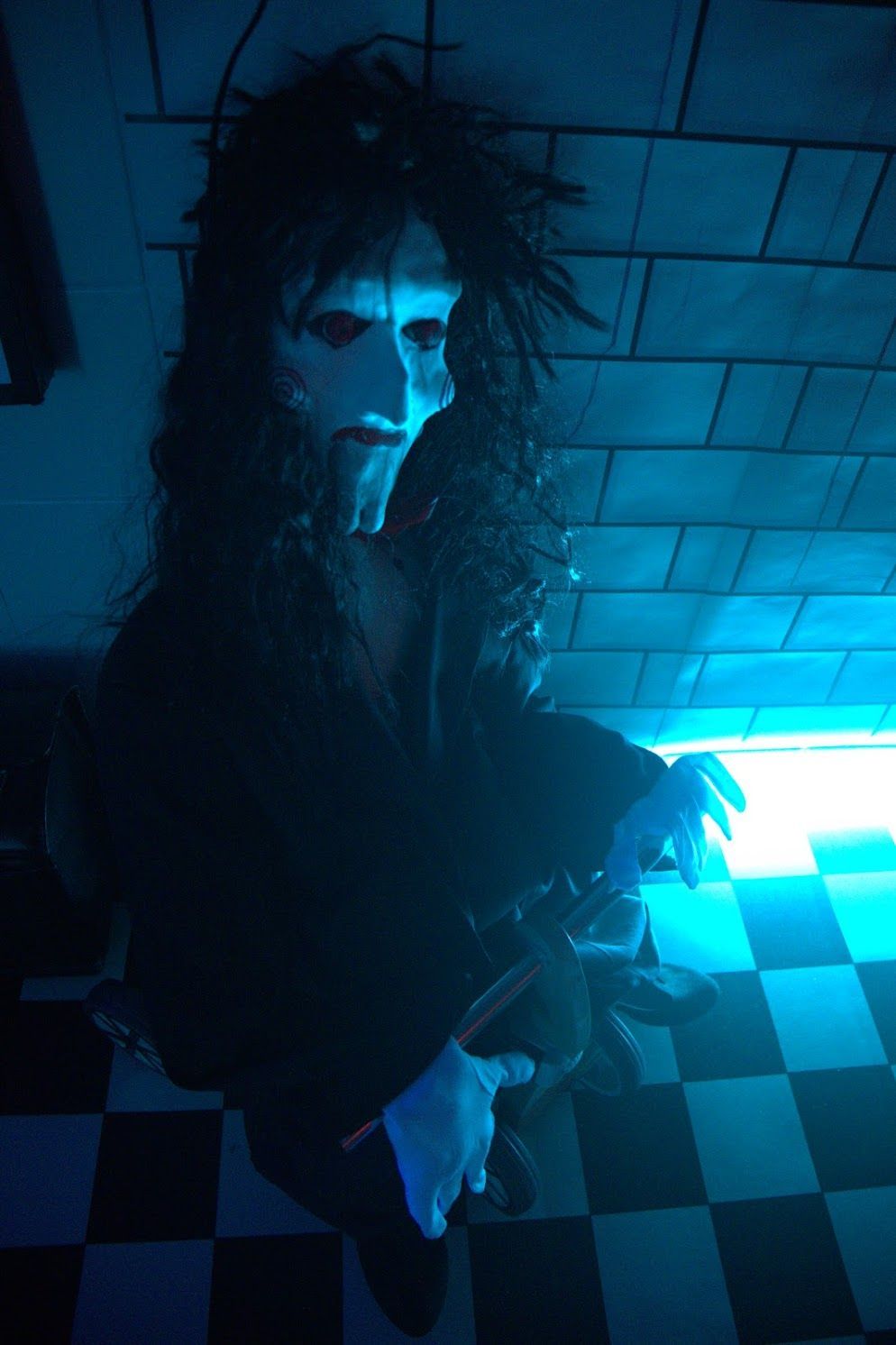 Gestalt mit weißer Maske und schwarzem Gewand vor kariertem Boden und blau beleuchteten Wänden.