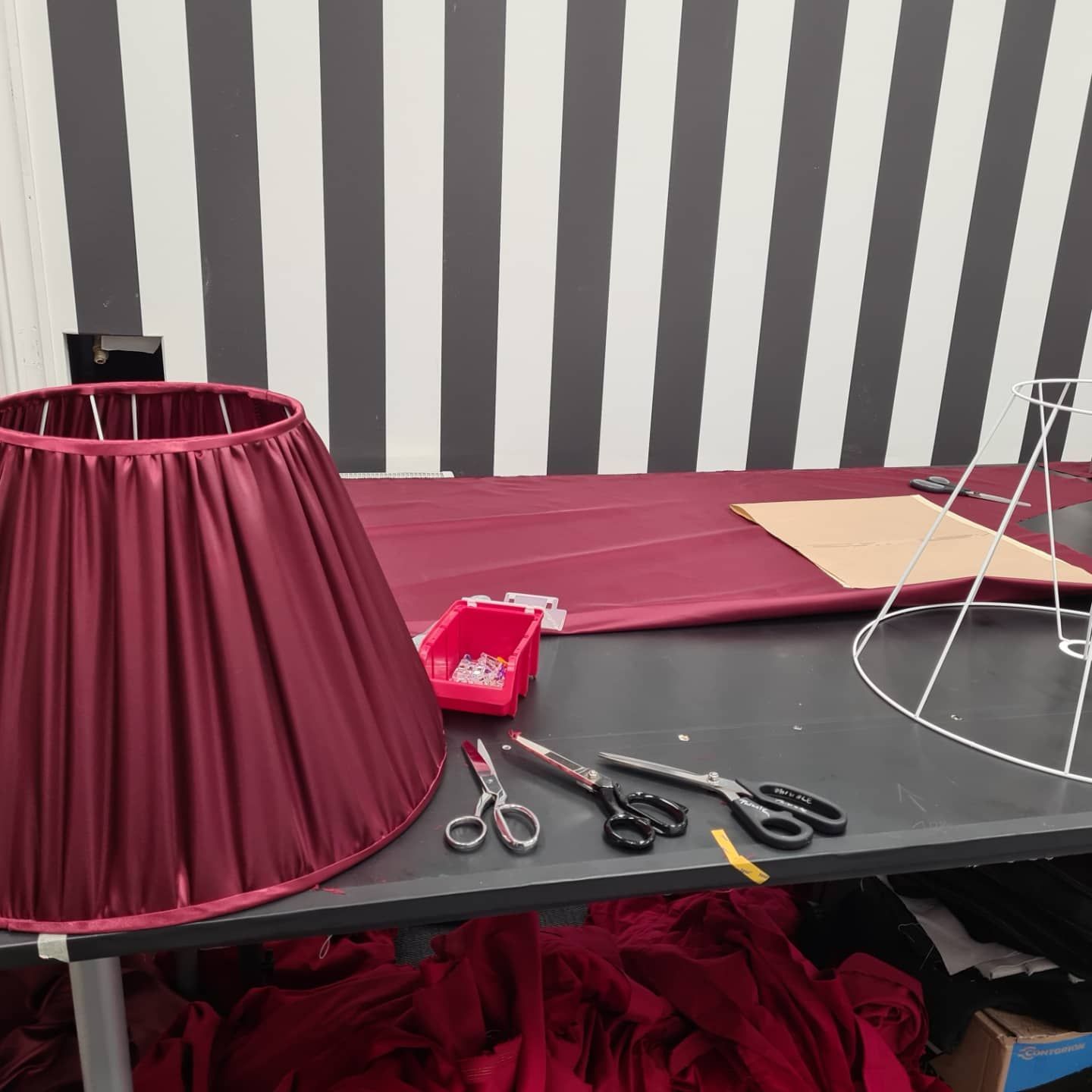 Auf einem Tisch wird mit Werkzeugen und Stoff ein Lampenschirm gefertigt. Burgunderrote Materialien.