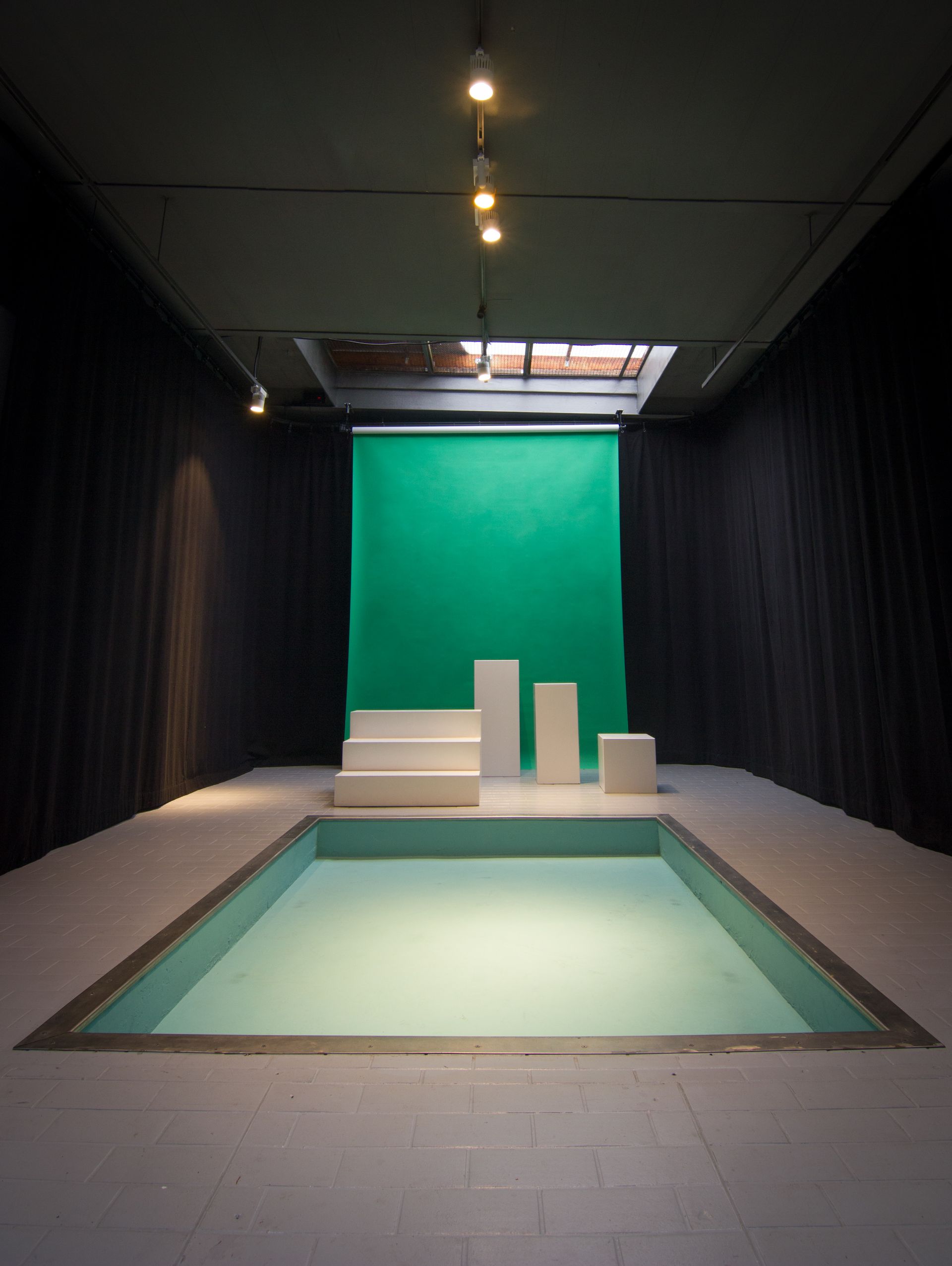 Indoor-Studio mit Pool, weißen Strukturen, grünem Hintergrund, schwarzen Vorhängen und Deckenbeleuchtung.