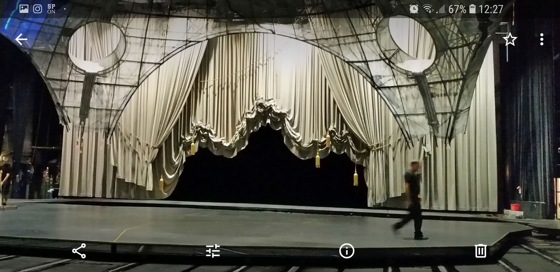 Eine Bühne mit einem Bühnenbild, eine Person, die über die Bühne geht, und ein reich verzierter Vorhang im Hintergrund.