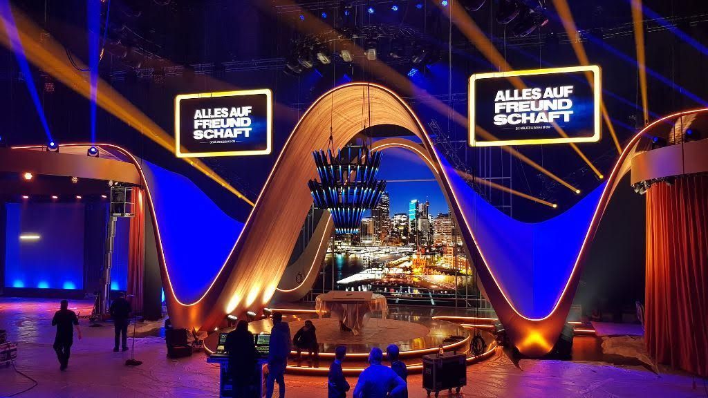 TV studio set with a large cityscape backdrop, stage lights, and the words 