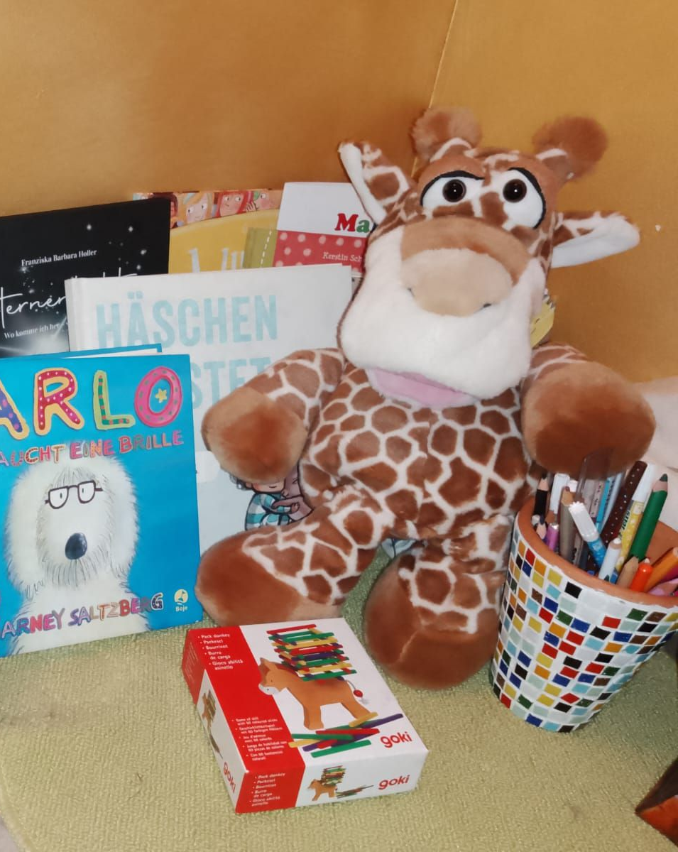 Foto von verschiedenen Kinderbüchern in einer Ecke. Davor sitzt eine Kuscheltier-Giraffe