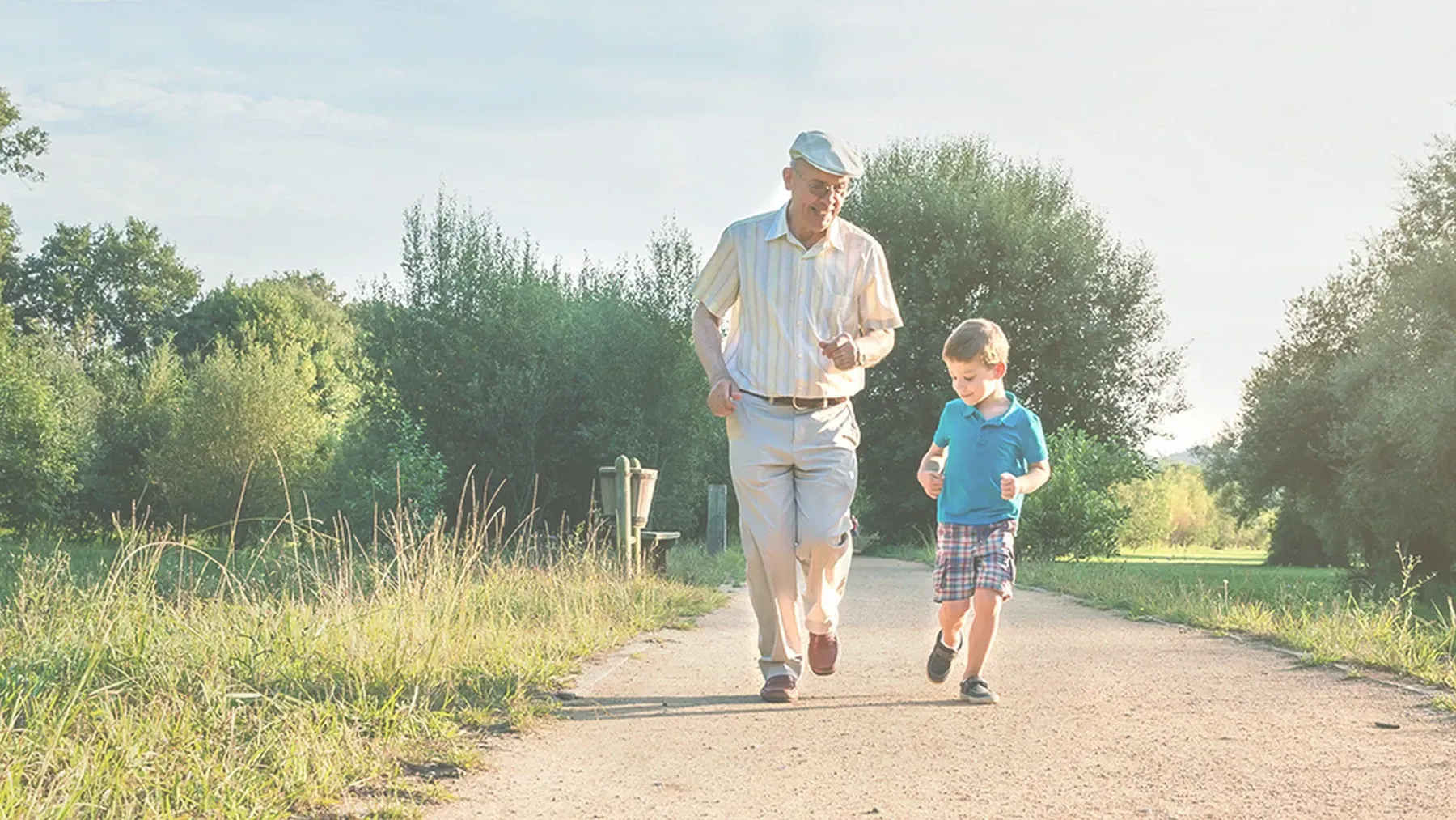 Eine Person und ein Kind joggen gemeinsam auf einem sonnenbeschienenen Kiesweg, der von Bäumen umgeben ist.