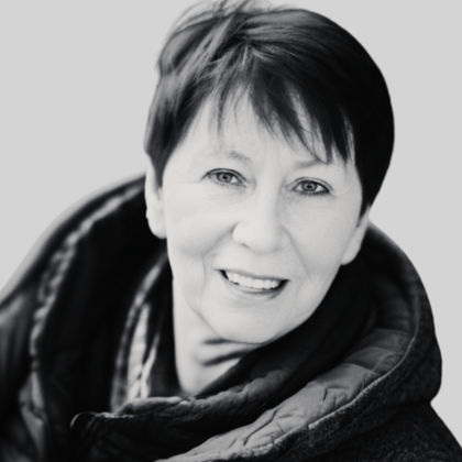 Frau mit kurzen dunklen Haaren lächelt, trägt eine Jacke vor hellem Hintergrund.