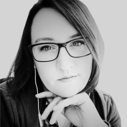 Frau mit Brille und Ohrhörern, das Kinn auf die Hand gestützt, schaut in die Kamera.