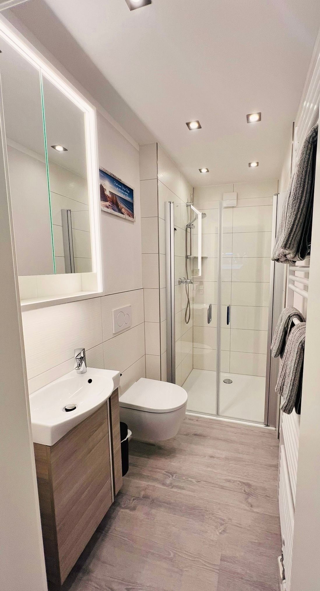 Ein helles, modernes Badezimmer mit Waschtisch aus Holz, wandhängender Toilette und verglaster, ebenerdiger Dusche.