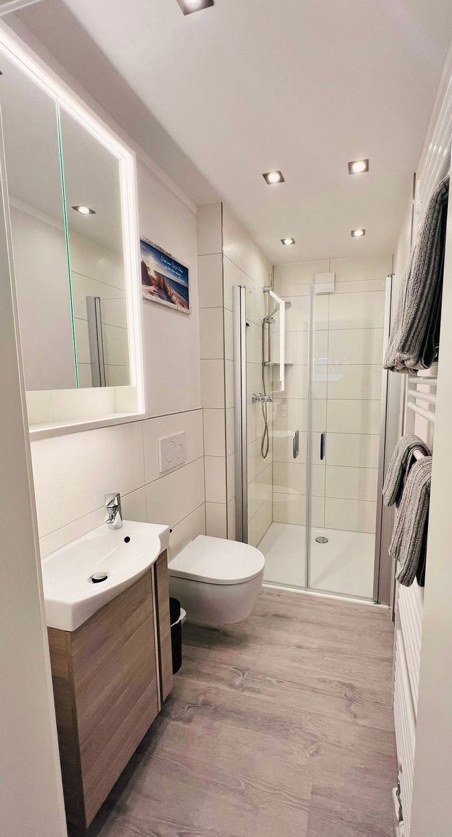 Ein helles, modernes Badezimmer mit Waschtisch aus Holz, wandhängender Toilette und verglaster, ebenerdiger Dusche.