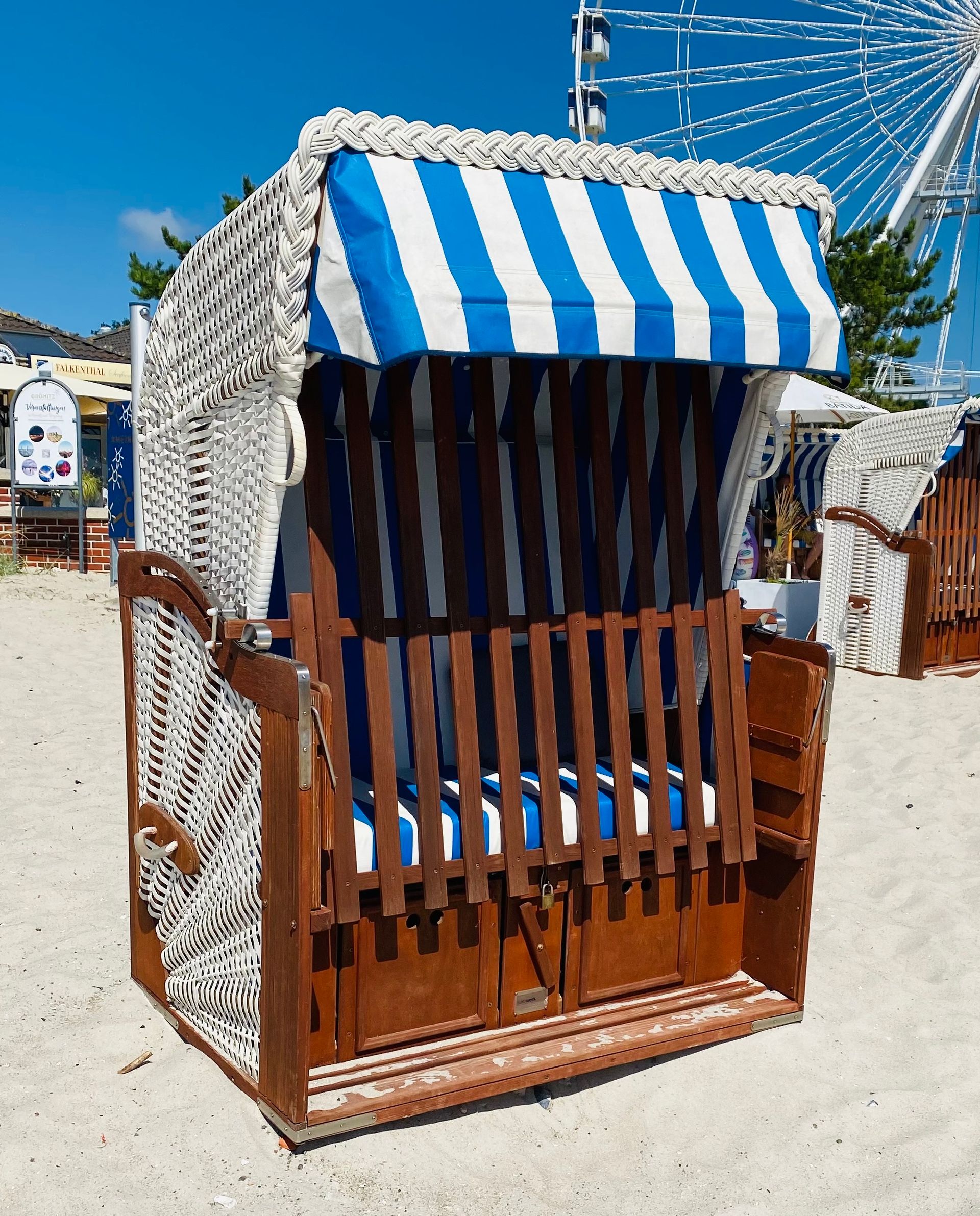 Ein klassischer blau-weiß gestreifter Strandkorb steht an einem Sandstrand, im Hintergrund ist ein Riesenrad zu sehen.