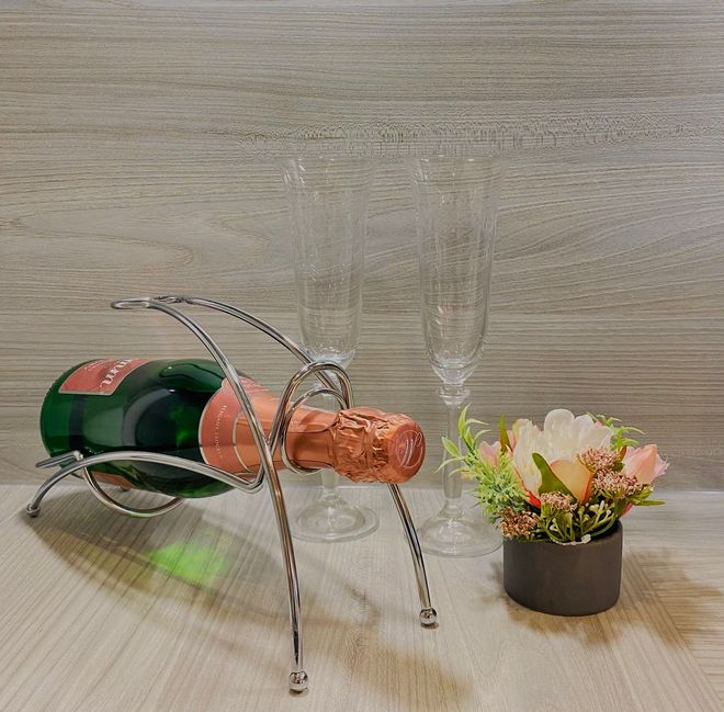 Eine Champagnerflasche in einem silbernen Drahtständer, daneben zwei leere Sektgläser und ein kleines Blumenarrangement in einem Topf auf einem Tisch.