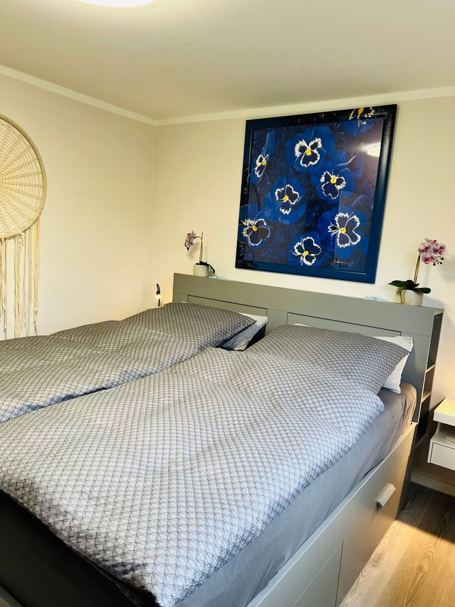 Ein Schlafzimmer mit einem Bett mit grau gemusterten Bettdecken, einem großen dunkelblauen floralen Wandbild und einem cremefarbenen Traumfänger.