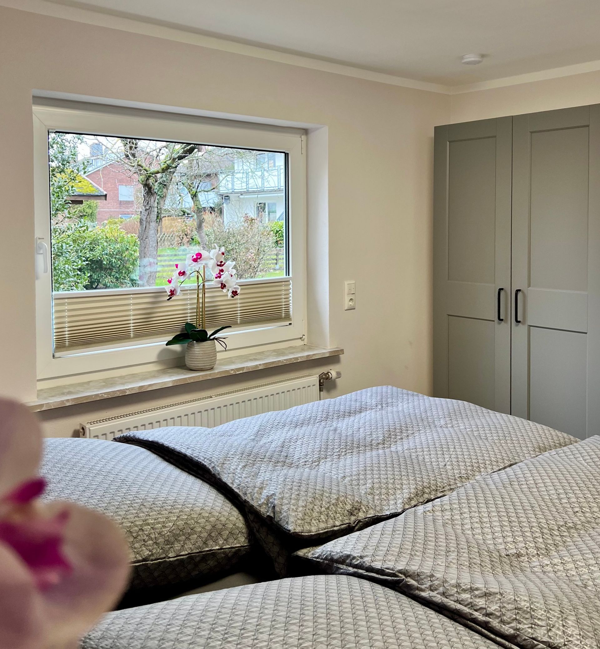Ein Schlafzimmer mit einem Bett mit strukturierter grauer Bettwäsche, einem großen Fenster, einer Orchidee im Topf und grauen Kleiderschranktüren.