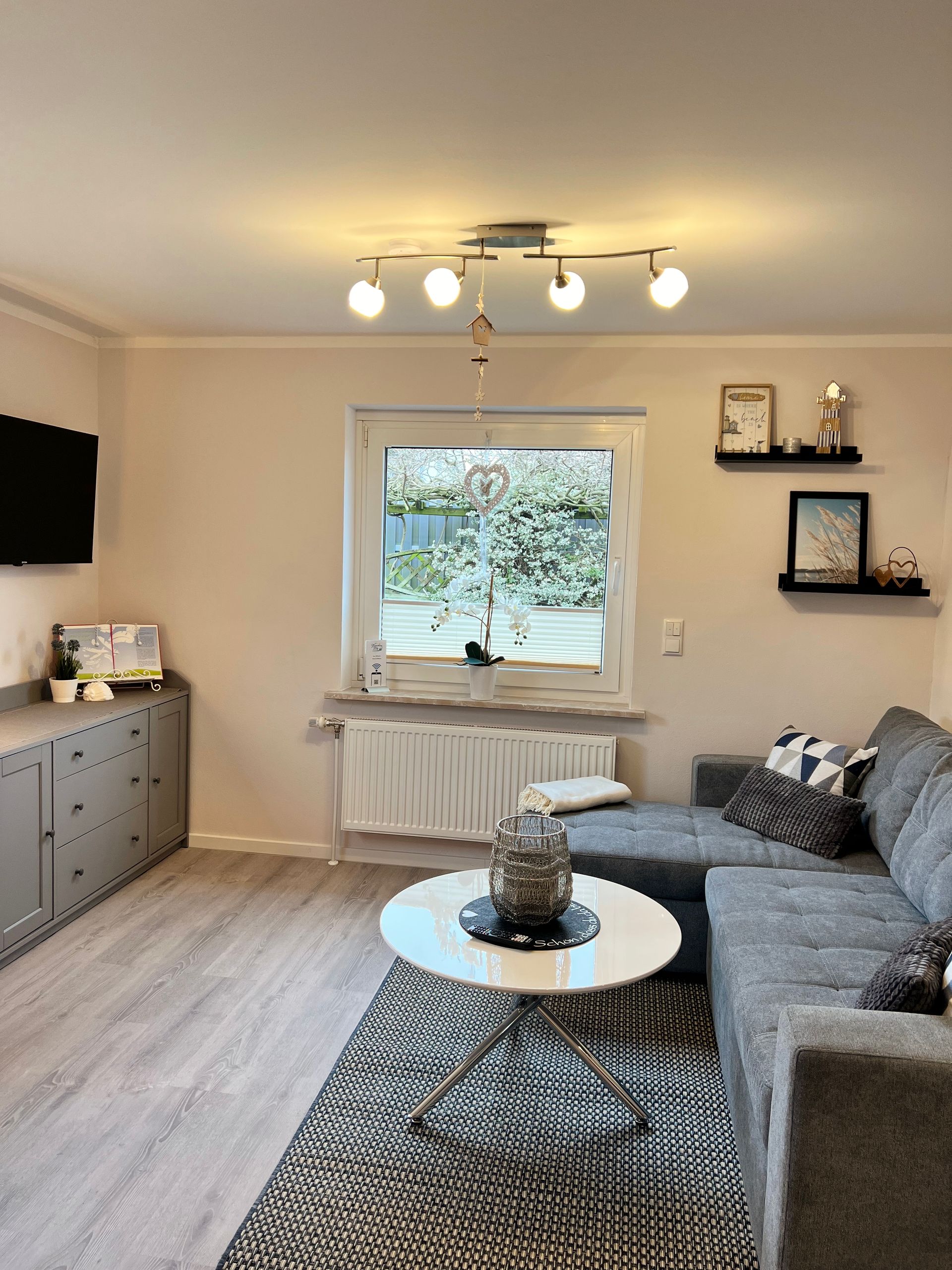 Ein Wohnzimmer mit einem grauen Ecksofa, einem weißen Couchtisch, einer grauen Kommode und zwei Wandregalen vor einer beigen Wand.