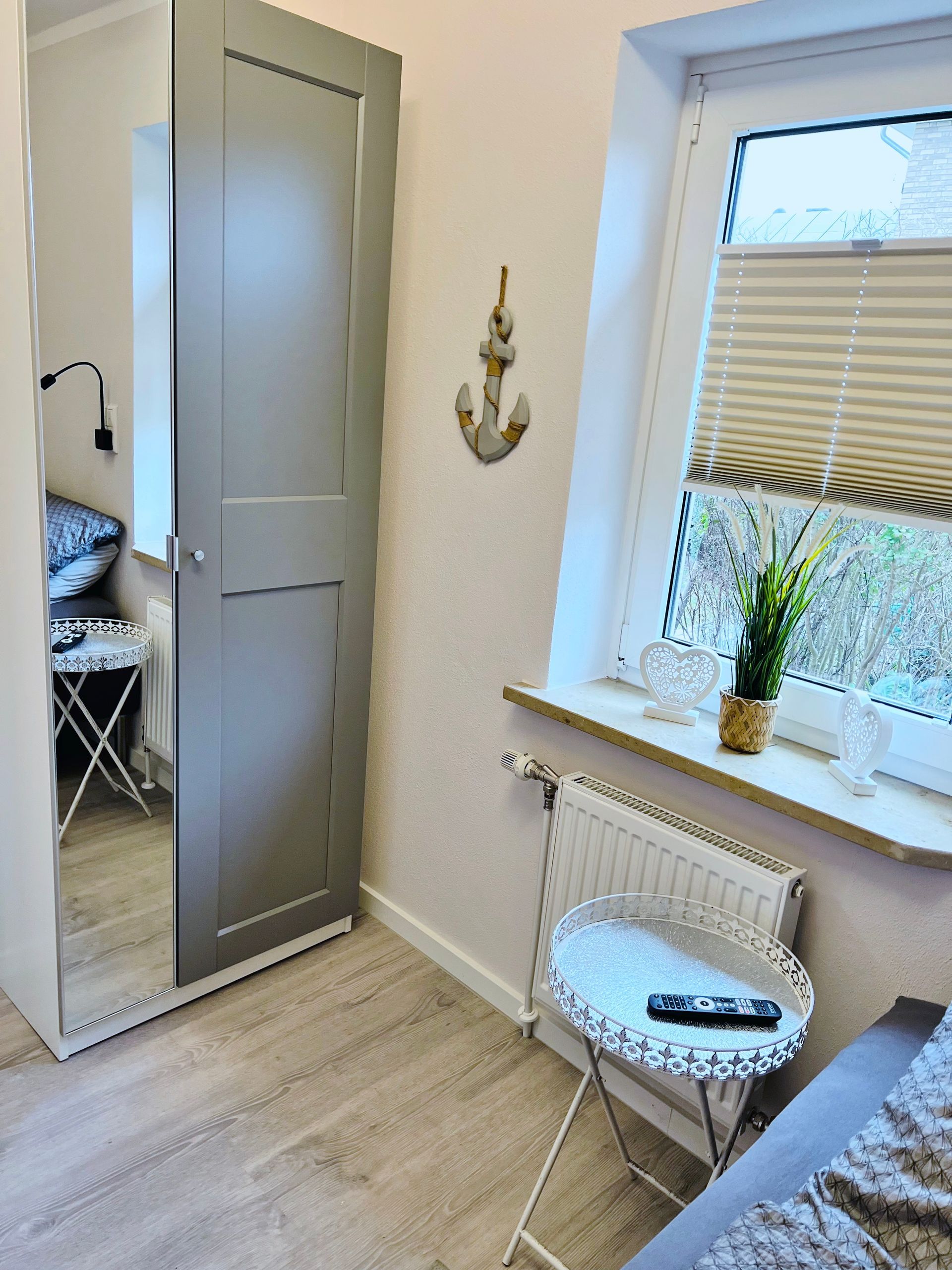 Ein Zimmer mit einem Kleiderschrank mit Spiegeltüren, einem Fenster mit Plissees, einer Topfpflanze und einem kleinen Beistelltisch aus Metall.
